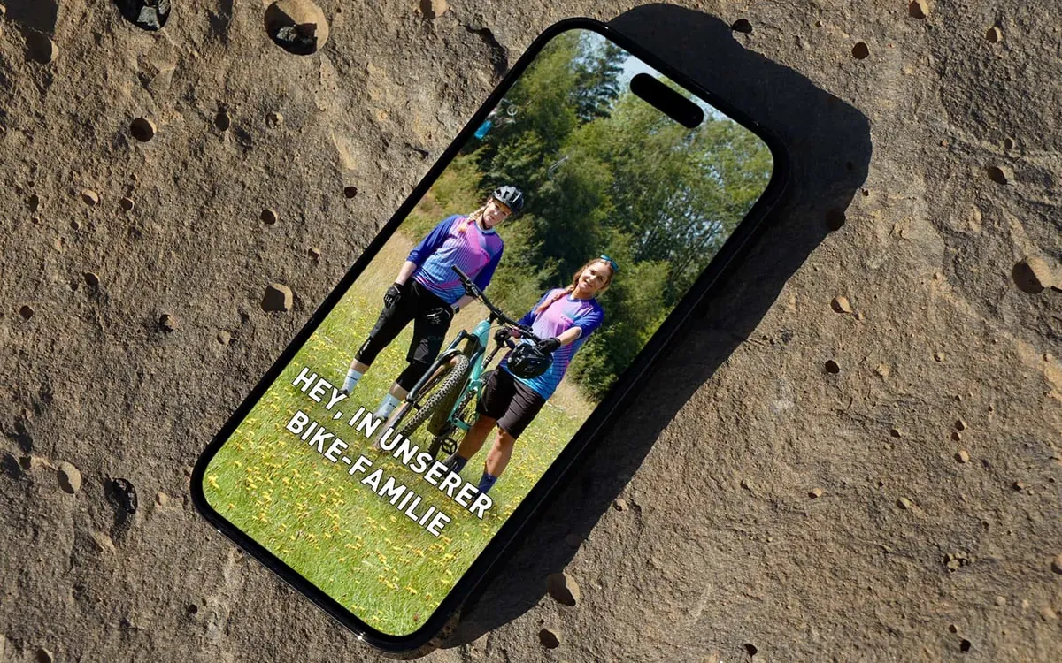 V&S Bike GmbH: Smartphone auf einem Felsen zeigt einen Social-Media-Beitrag mit zwei Radfahrerinnen und der Zeile „Hey in unserer Bike-Familie"