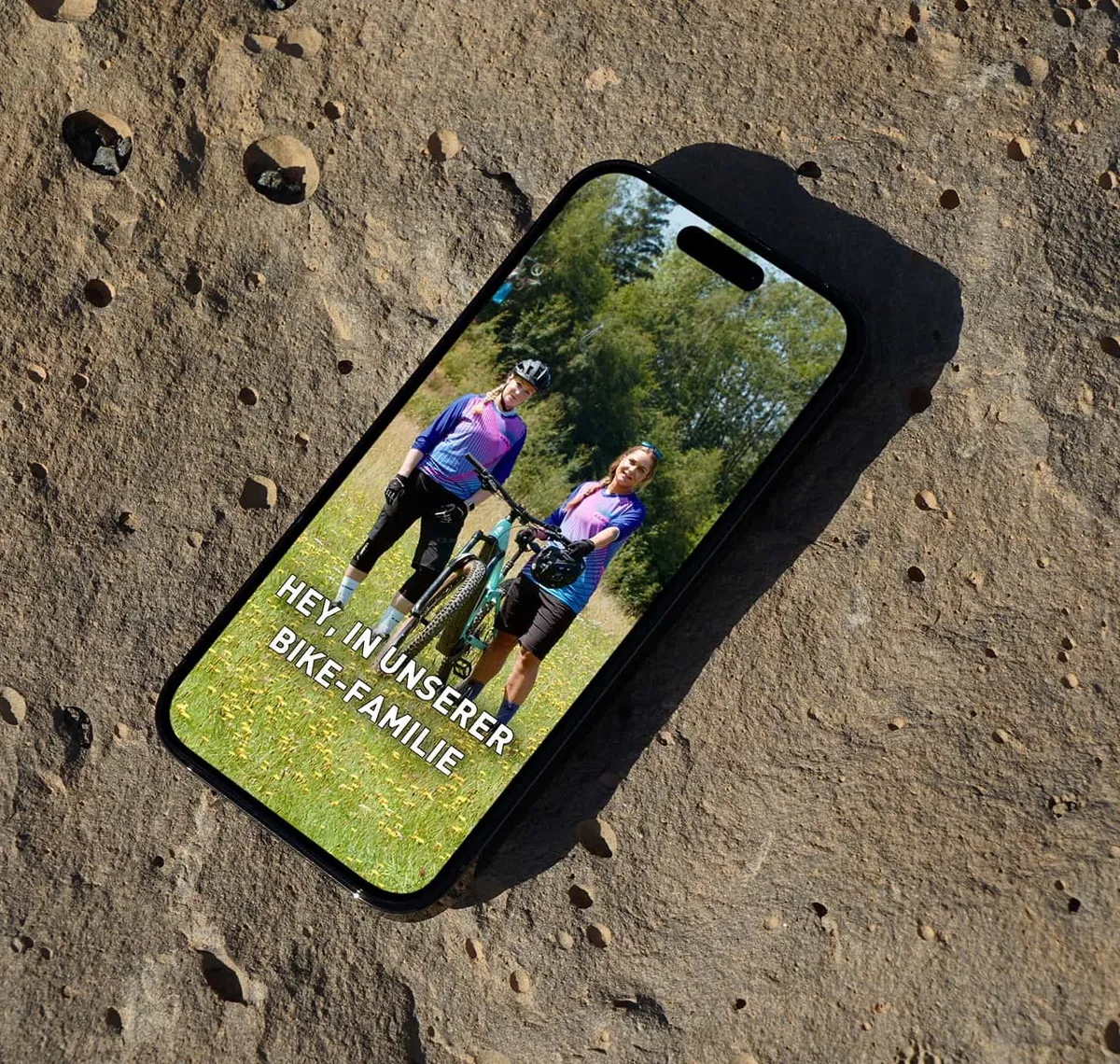 V&S Bike GmbH: Smartphone auf einem Felsen zeigt einen Social-Media-Beitrag mit zwei Radfahrerinnen und der Zeile „Hey in unserer Bike-Familie"