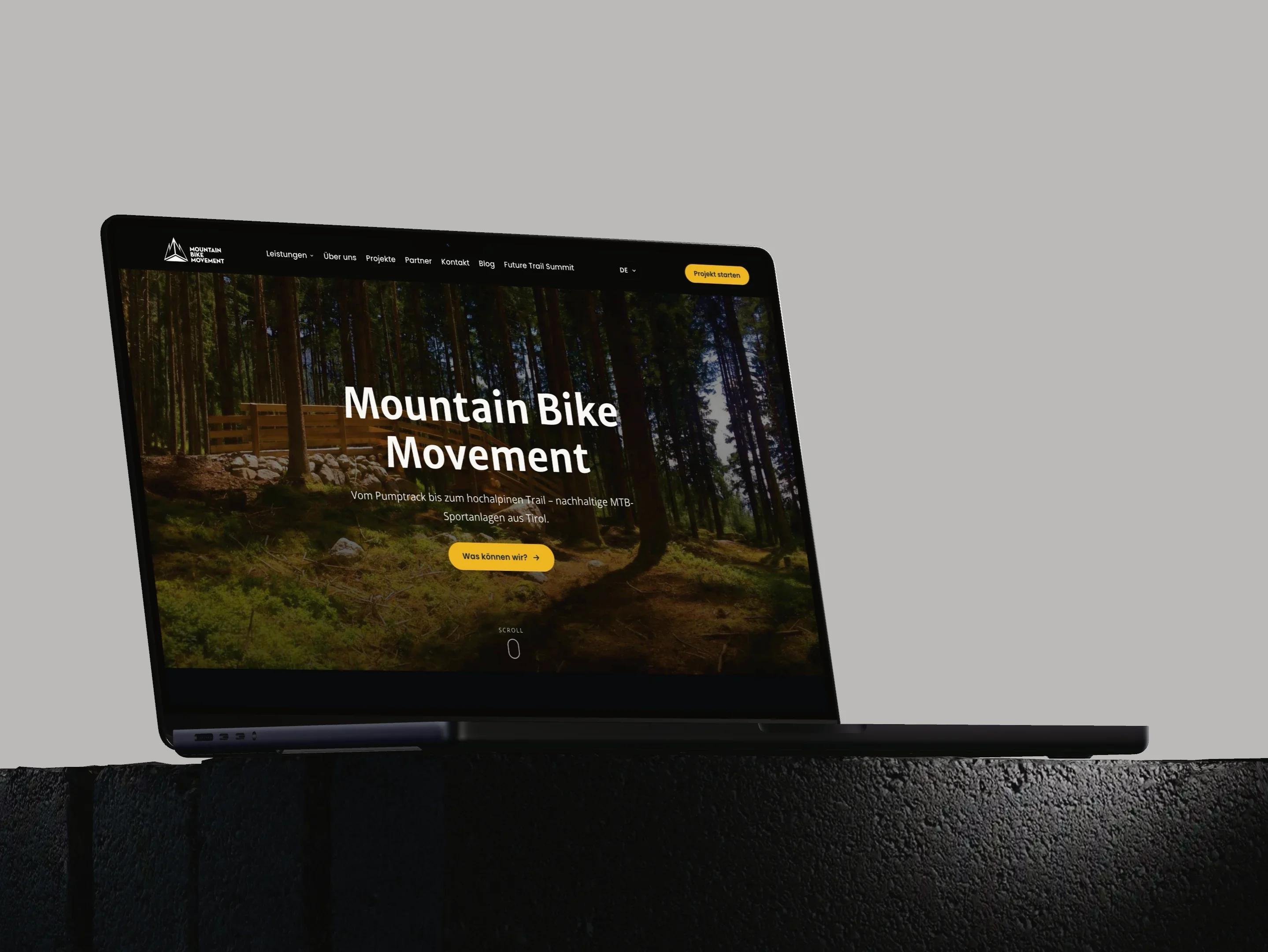 Mountain Bike Movement: Ein MacBook Pro auf einem dunklen Steinsockel vor hellgrauem Grund zeigt die Startseite von mountainbikemovement.com mit einem Waldtrail und dem Schriftzug Mountain Bike Movement.