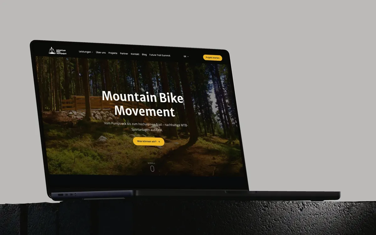 Mountain Bike Movement: Ein MacBook Pro auf einem dunklen Steinsockel vor hellgrauem Grund zeigt die Startseite von mountainbikemovement.com mit einem Waldtrail und dem Schriftzug Mountain Bike Movement.