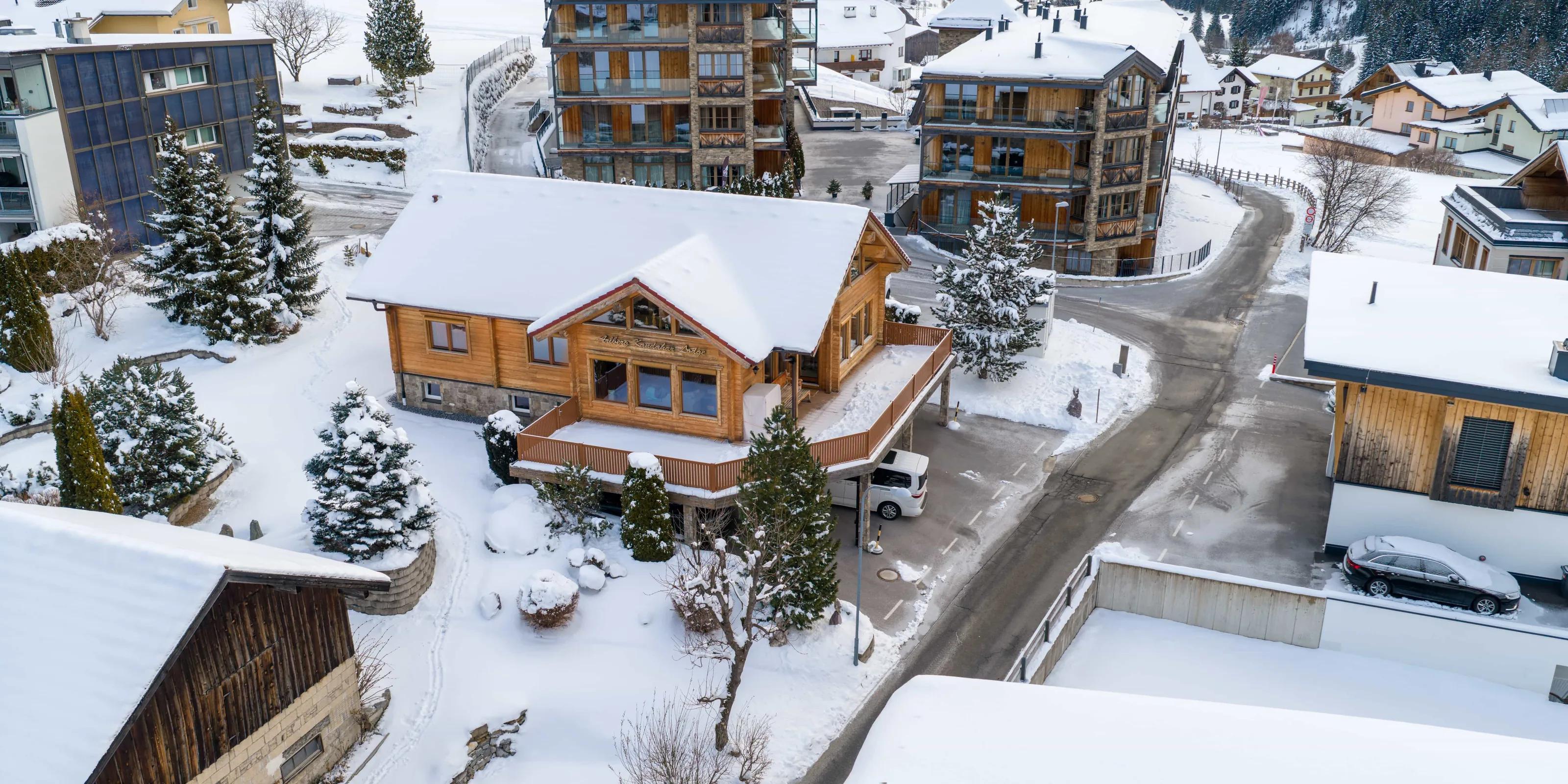 Virtueller Rundgang: Die Arlberg Kandahar Lodge aus der Luft im Winter: das verschneite Holzhaus zwischen den Nachbarhäusern von St. Anton, dahinter der Hang