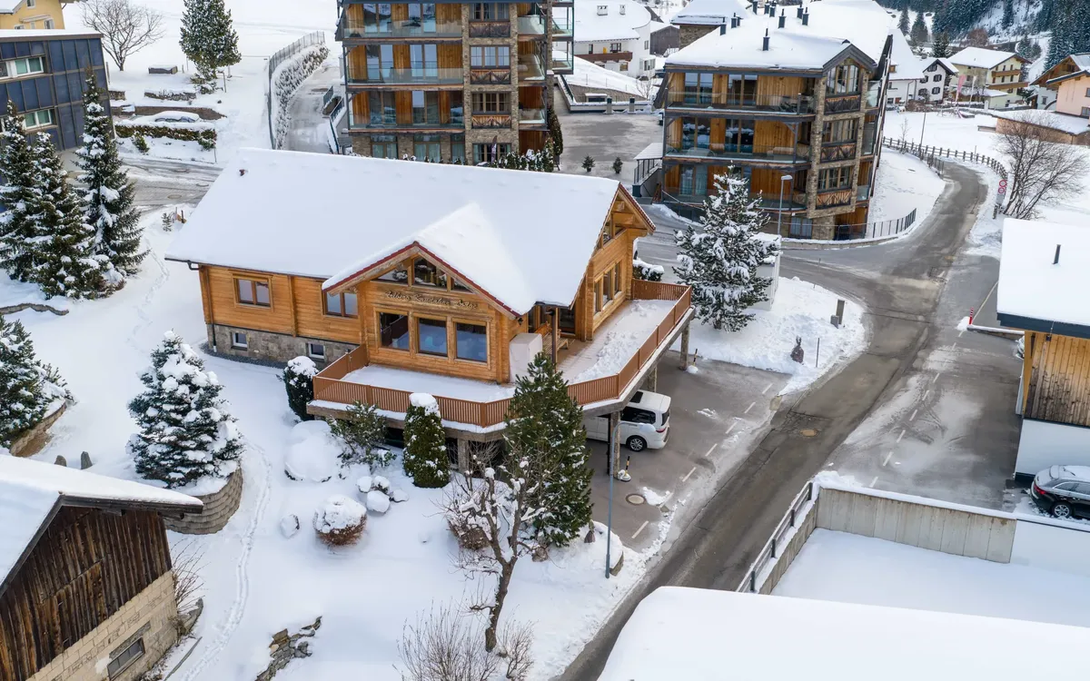 Virtueller Rundgang: Die Arlberg Kandahar Lodge aus der Luft im Winter: das verschneite Holzhaus zwischen den Nachbarhäusern von St. Anton, dahinter der Hang