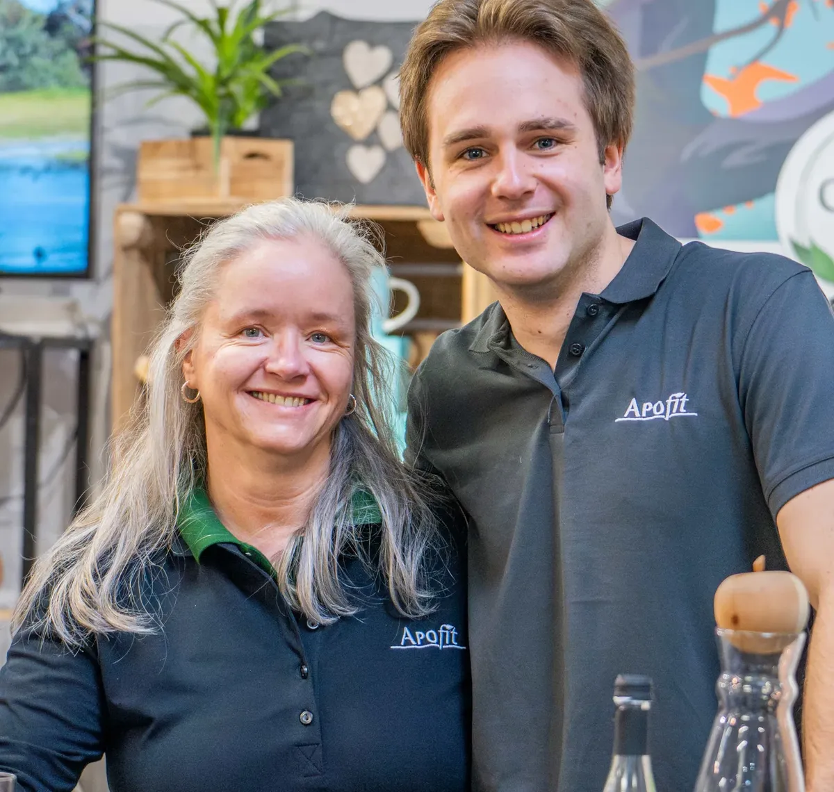 APOfit: Eine Mitarbeiterin und ein Mitarbeiter von APOfit hinter der Theke am Messestand, dahinter ein Roll-up mit der Zeile Heilen mit der Natur und ein Bildschirm mit dem Film.