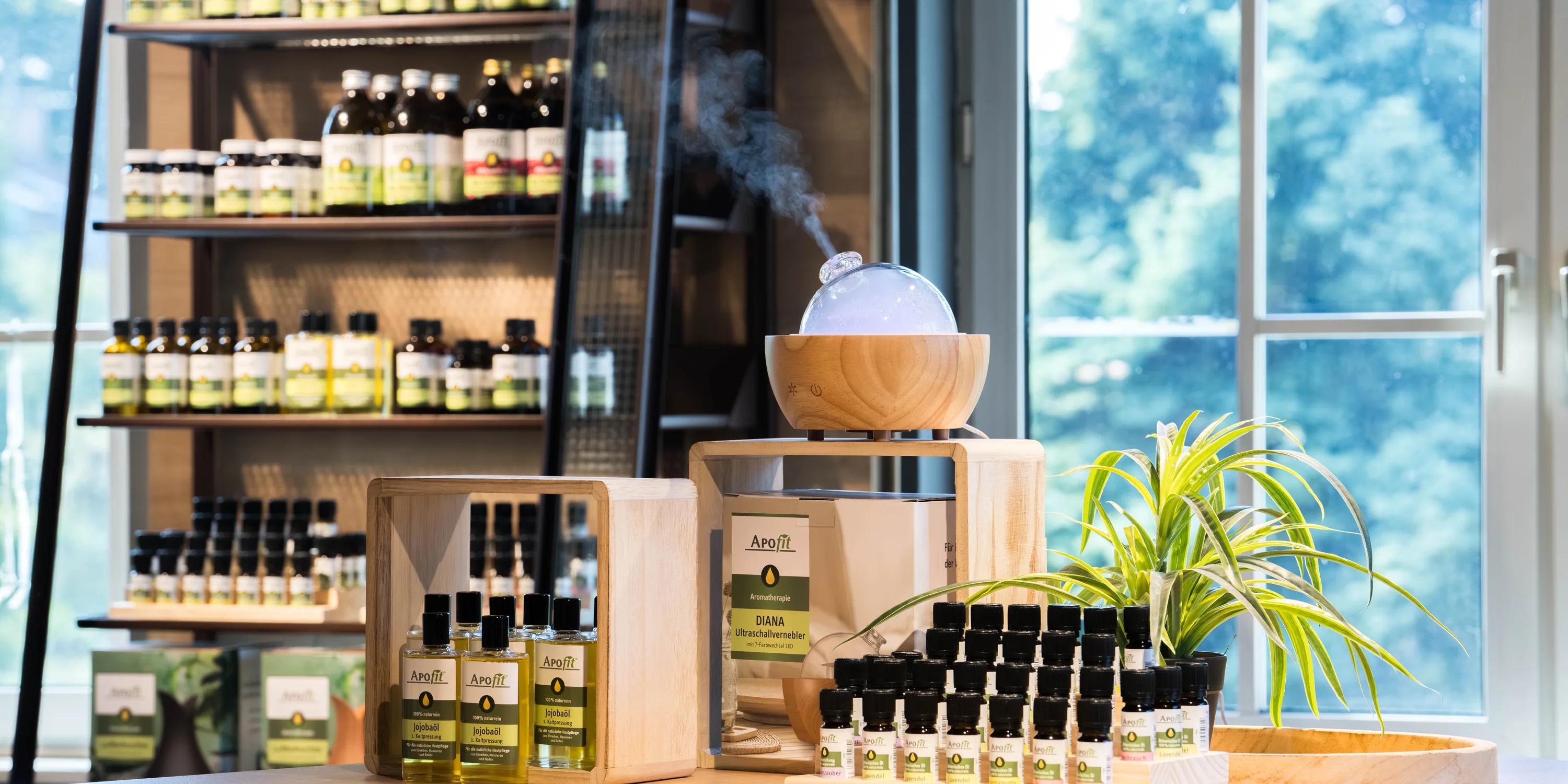 APOfit: Ein Regal mit APOfit-Produkten, davor auf einem Schreibtisch eine Holzbox mit ätherischen Ölen und ein Diffuser aus Holz, aus dem Dampf steigt, dahinter ein grosses Fenster.