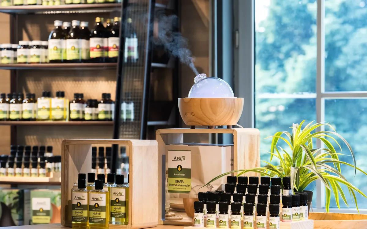 APOfit: Ein Regal mit APOfit-Produkten, davor auf einem Schreibtisch eine Holzbox mit ätherischen Ölen und ein Diffuser aus Holz, aus dem Dampf steigt, dahinter ein grosses Fenster.