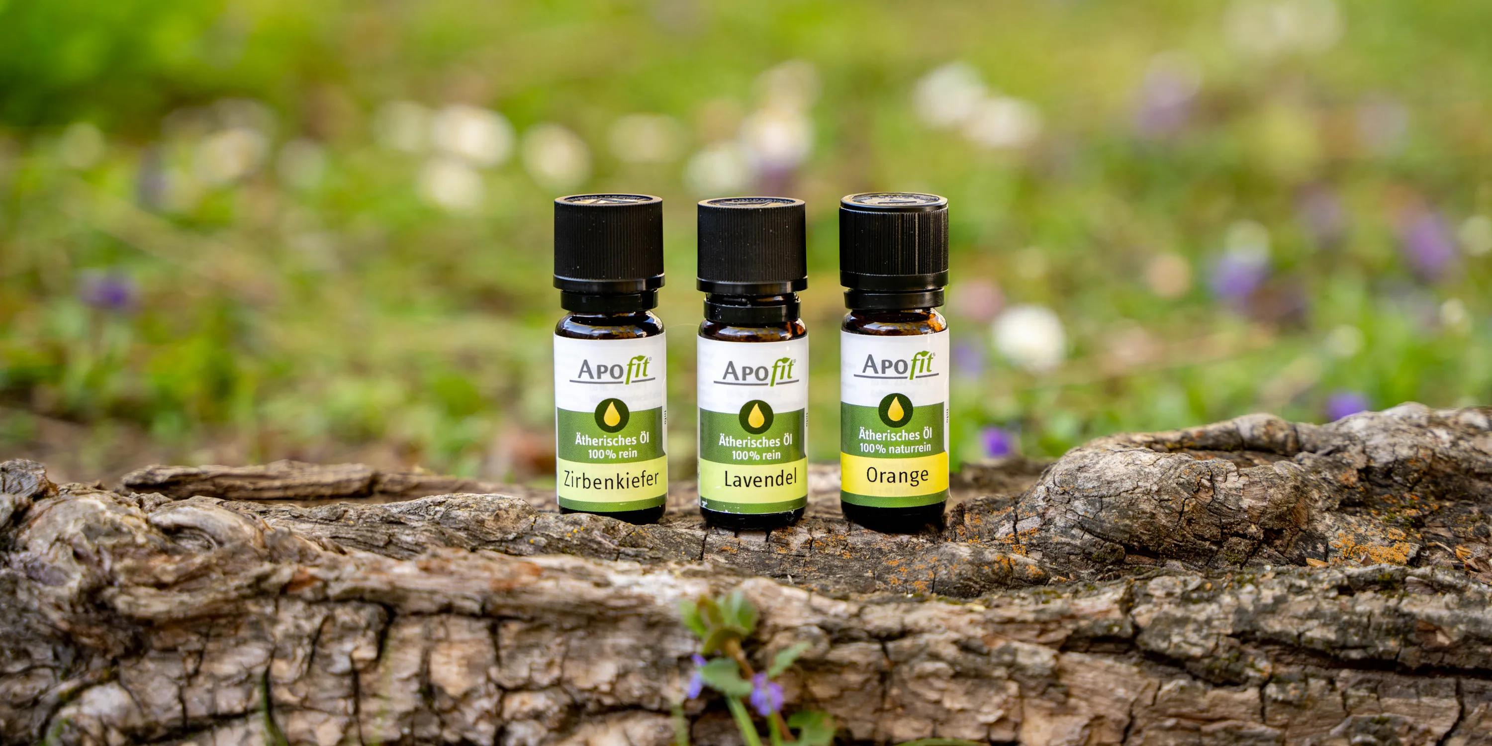 Produktfotos für APOfit: Drei Fläschchen APOfit Ätherisches Öl, Zirbenkiefer, Lavendel und Orange, stehen auf einer Baumrinde vor einer Wiese mit Frühlingsblumen.