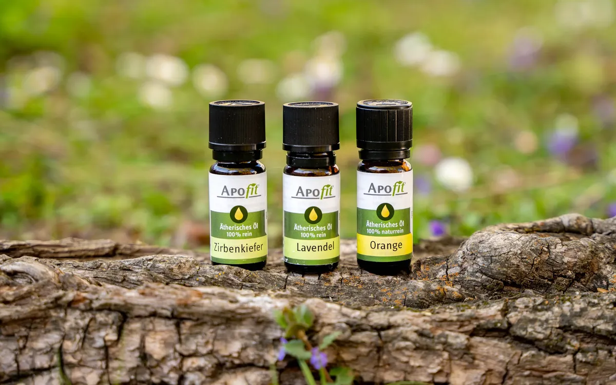 Produktfotos für APOfit: Drei Fläschchen APOfit Ätherisches Öl, Zirbenkiefer, Lavendel und Orange, stehen auf einer Baumrinde vor einer Wiese mit Frühlingsblumen.