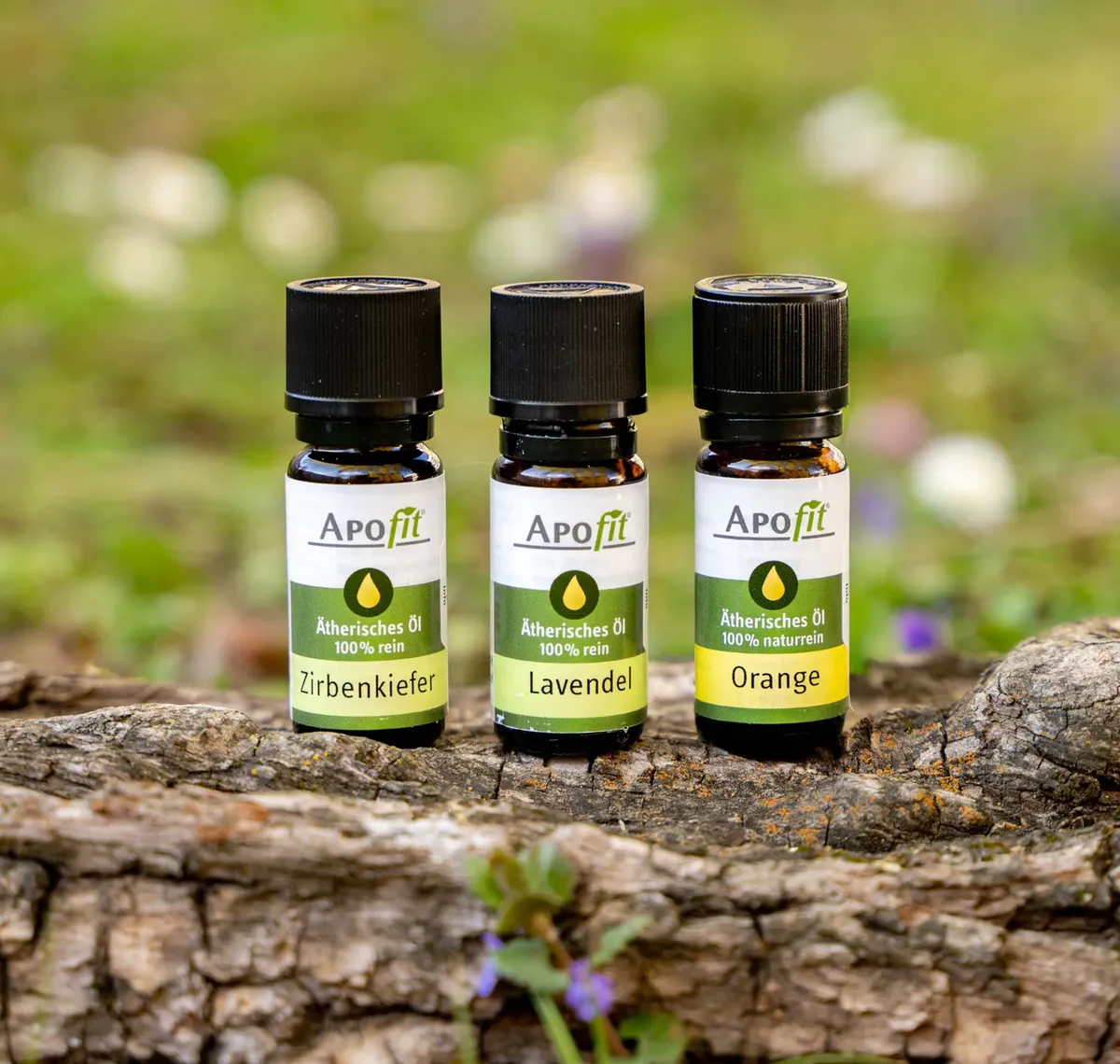 APOfit: Drei Fläschchen APOfit Ätherisches Öl, Zirbenkiefer, Lavendel und Orange, stehen auf einer Baumrinde vor einer Wiese mit Frühlingsblumen.