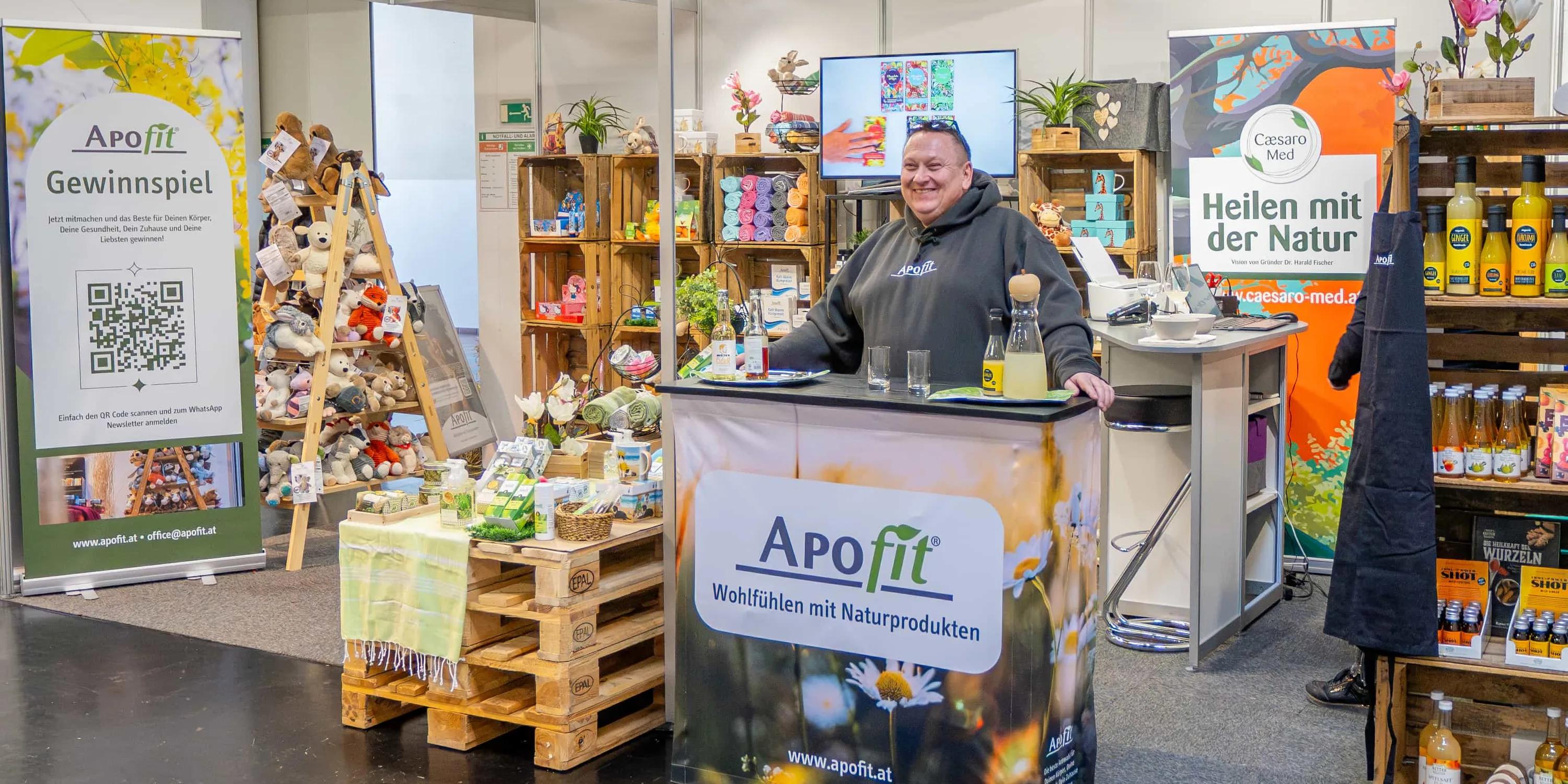 APOfit: Der Messestand von APOfit: ein Roll-up, Holzkisten mit Produkten, die Theke mit dem Wiesenmotiv und dem Logo, darüber das Schild mit dem Namen.