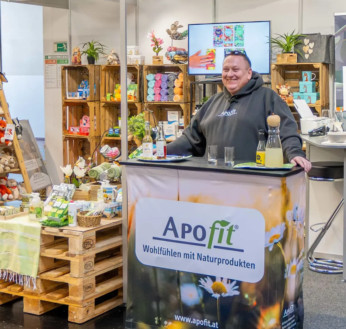 APOfit: Der Messestand von APOfit: ein Roll-up, Holzkisten mit Produkten, die Theke mit dem Wiesenmotiv und dem Logo, darüber das Schild mit dem Namen.