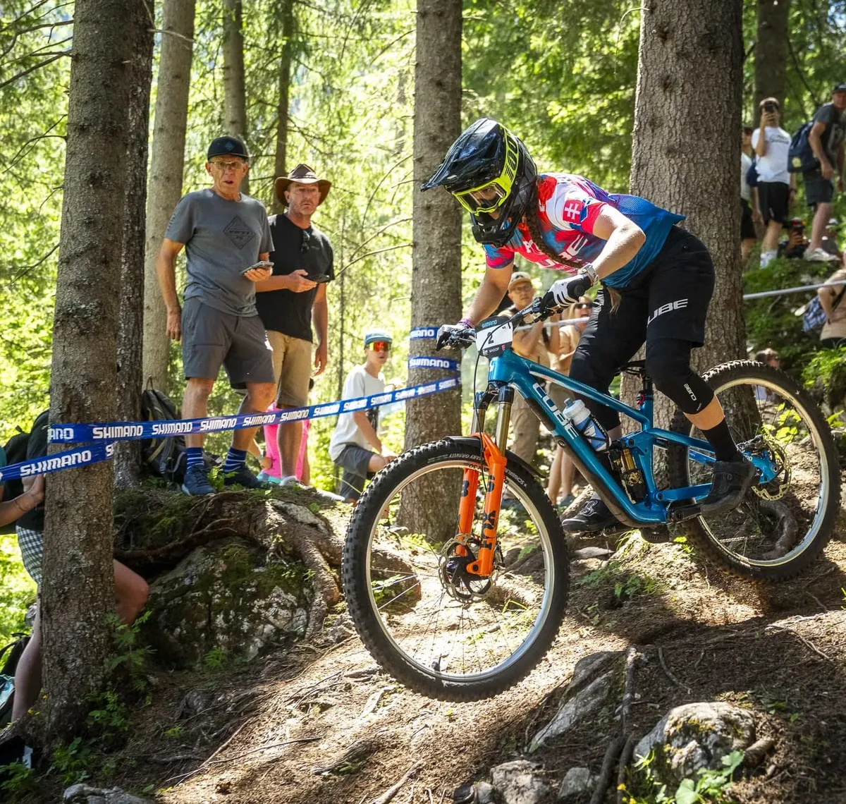 CUBE Actionteam: Downhill-Fahrer im Rennen auf einem Wurzeltrail, Zuschauer hinter dem Absperrband