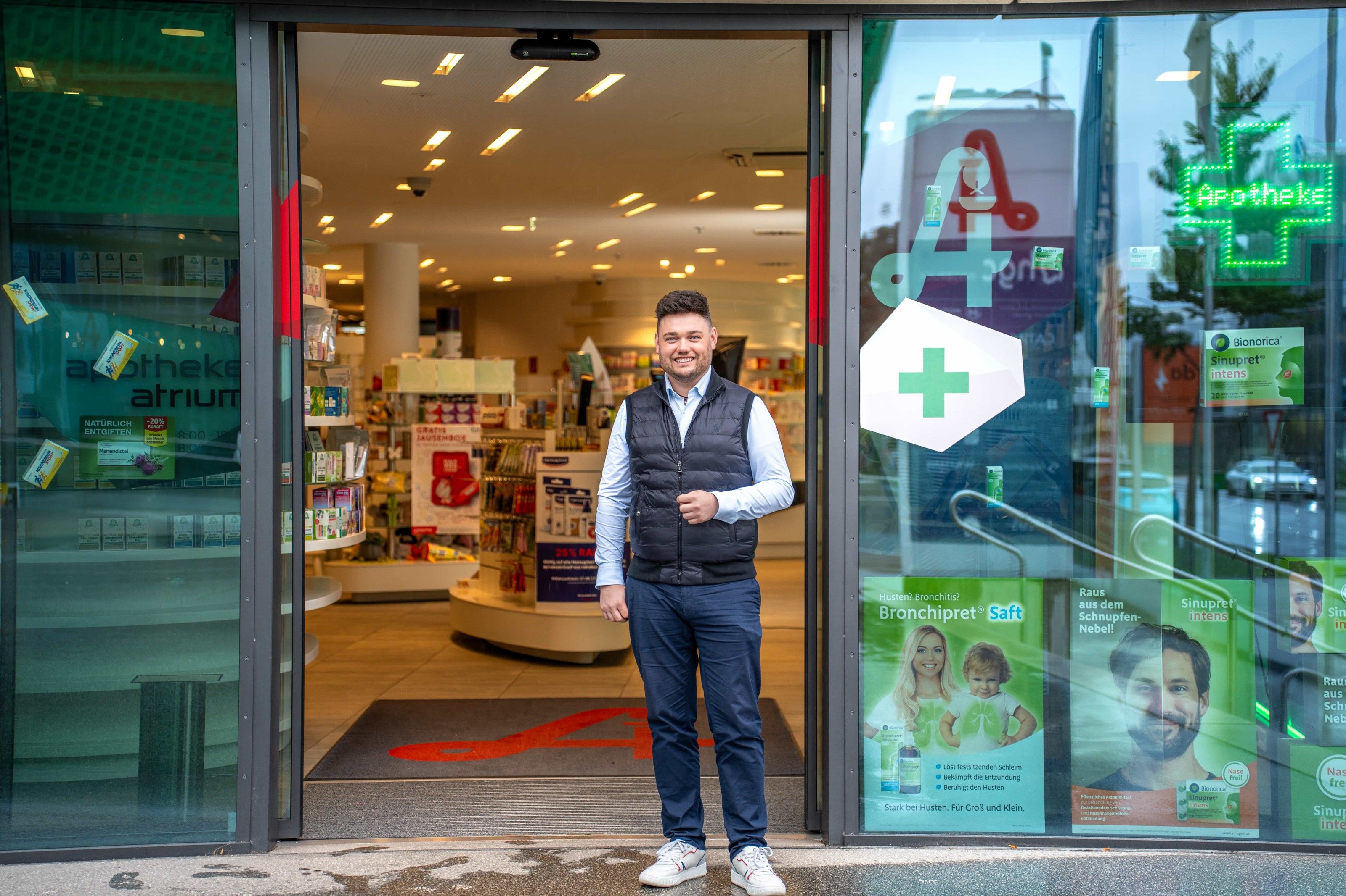 provivamed: Mitarbeiter steht im geöffneten Eingang einer Apotheke, rechts das grüne Apothekenkreuz