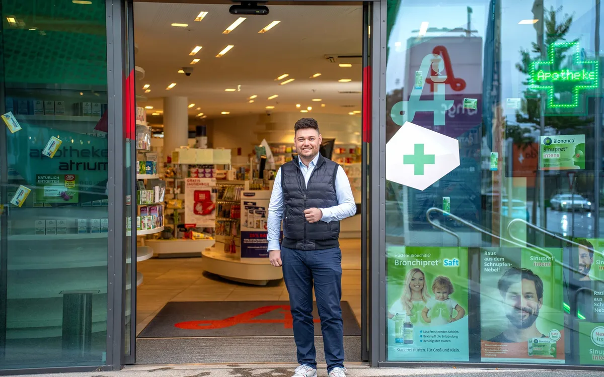 provivamed: Mitarbeiter steht im geöffneten Eingang einer Apotheke, rechts das grüne Apothekenkreuz
