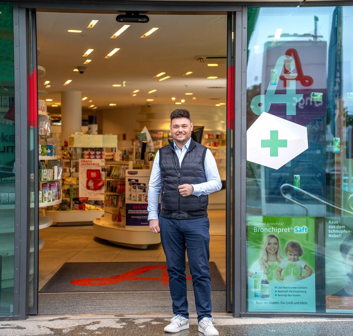 provivamed: Mitarbeiter steht im geöffneten Eingang einer Apotheke, rechts das grüne Apothekenkreuz