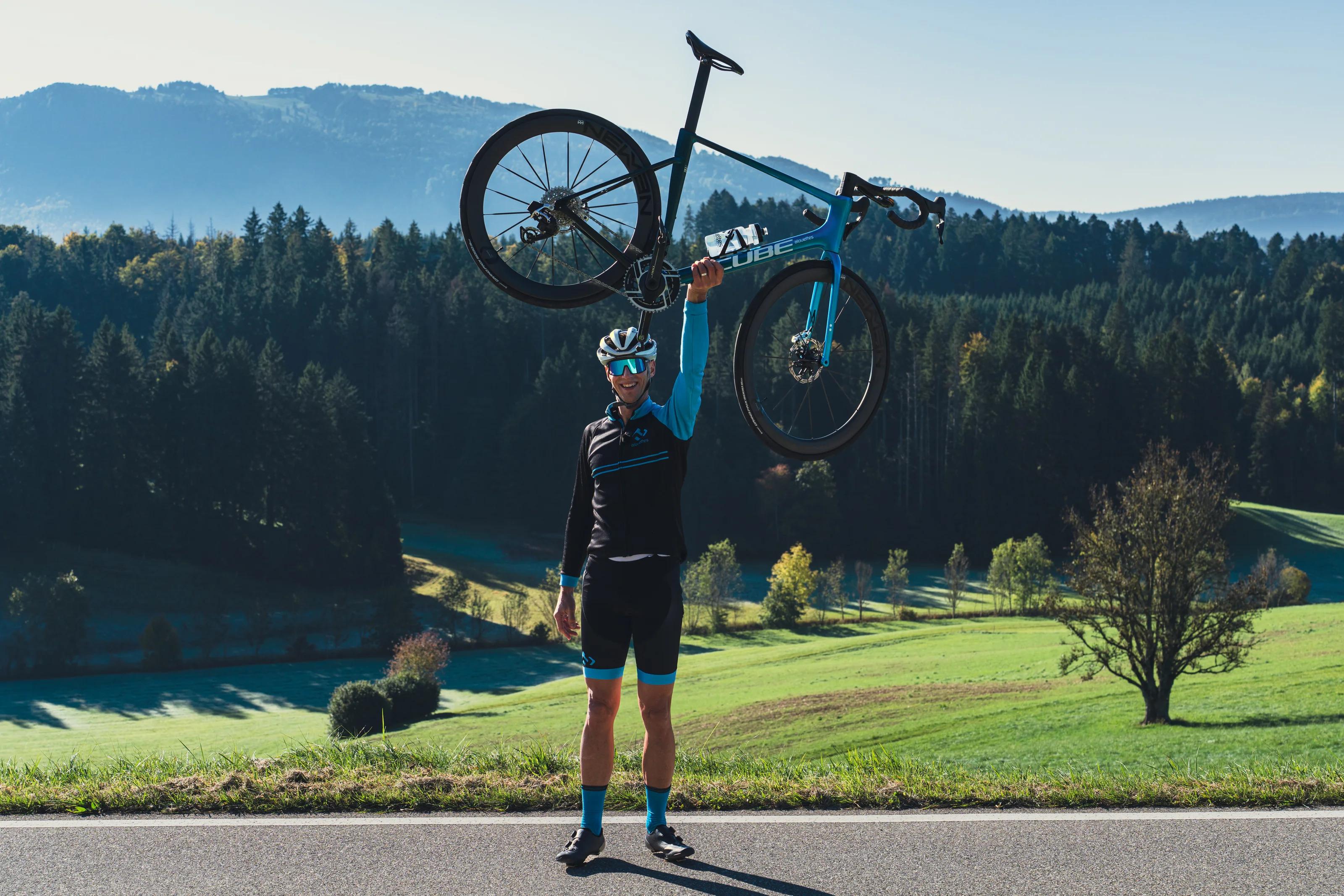 CUBE Store Valbirse: Ein Fahrer hebt sein Rennrad über den Kopf, hinter ihm der bewaldete Jura.