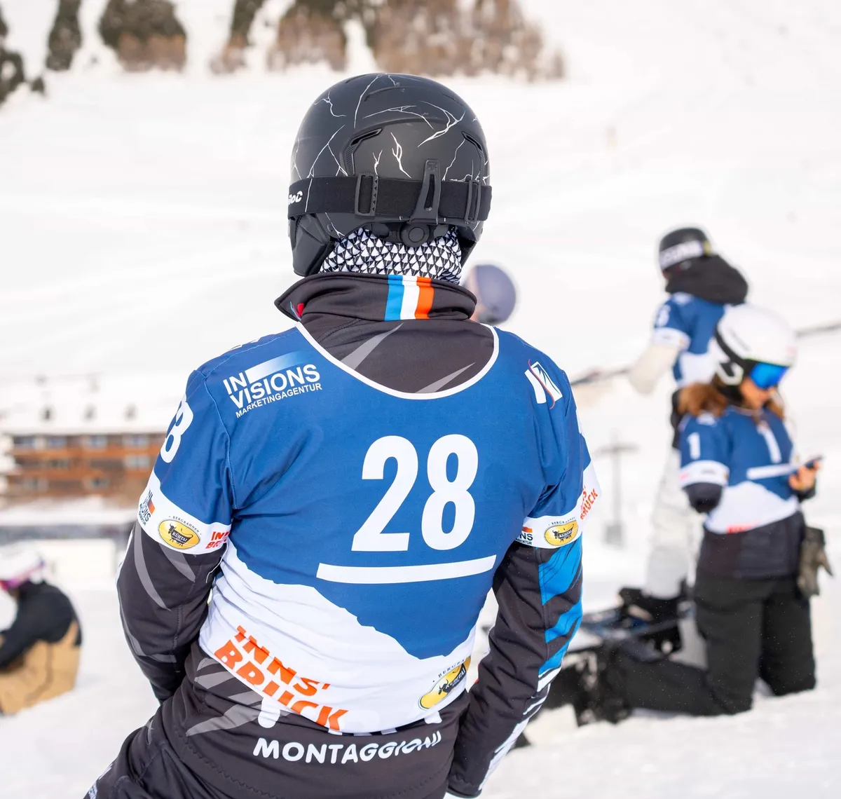 Crazy Curves / FIS Para Snowboard Worldcup: Rennläufer von hinten mit der Startnummer 28 und dem Invisions-Logo auf dem Trikot, dahinter ein Kamerateam im Schnee