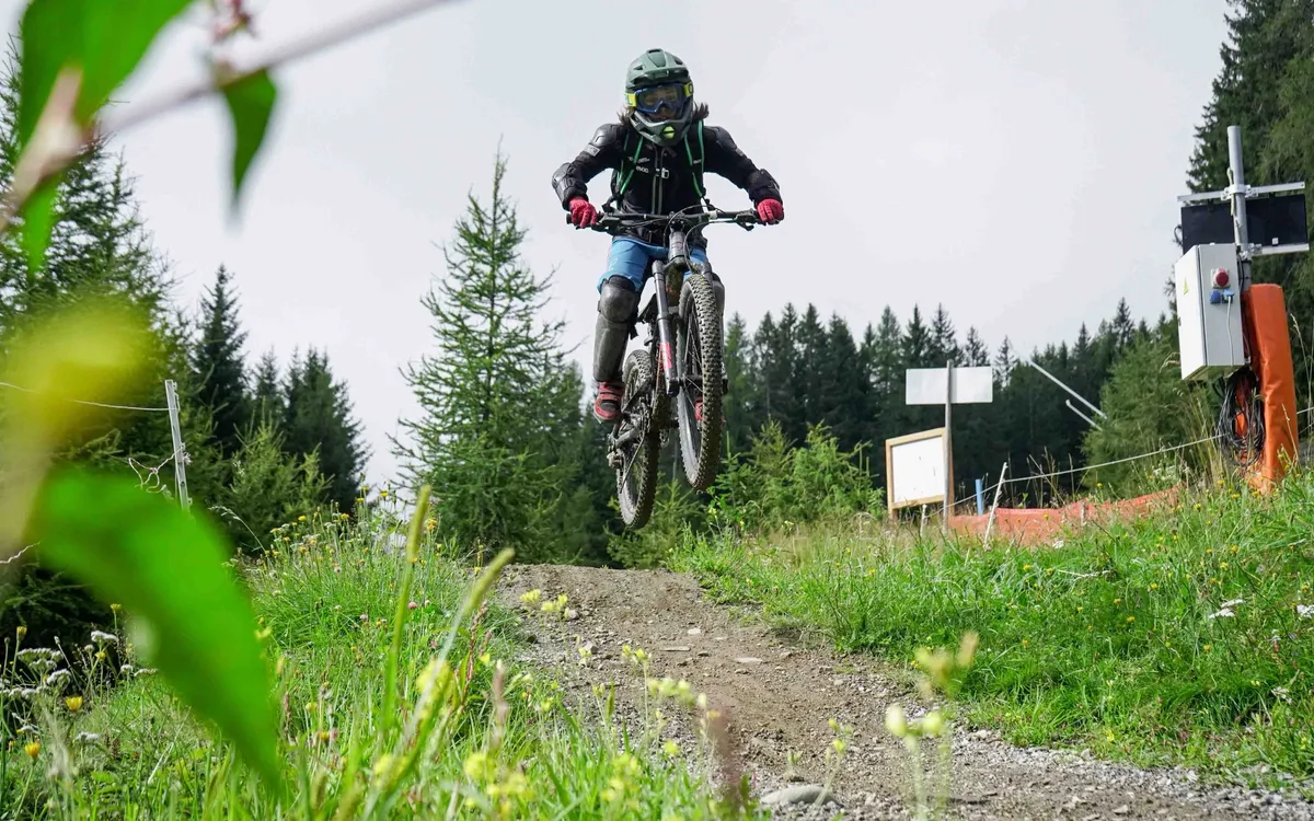 MTB & Downhill Verein Tirol: Downhill-Fahrer springt über eine Kuppe der Bikepark-Strecke, dahinter Nadelwald