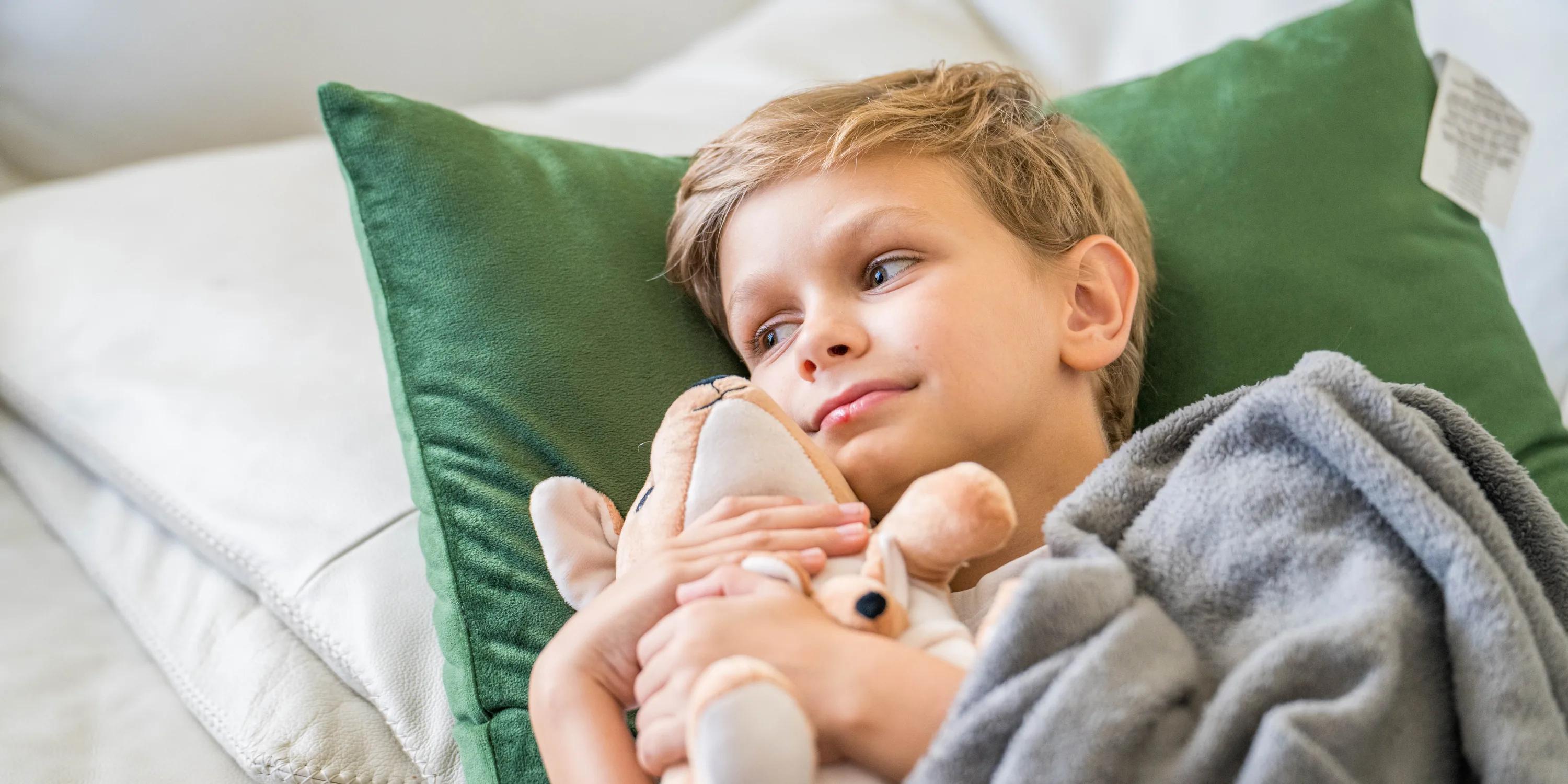 Social Media für APOfit: Ein Bub liegt mit seinem Warmie-Plüschtier auf einem grünen Kissen, zugedeckt mit einer grauen Decke.
