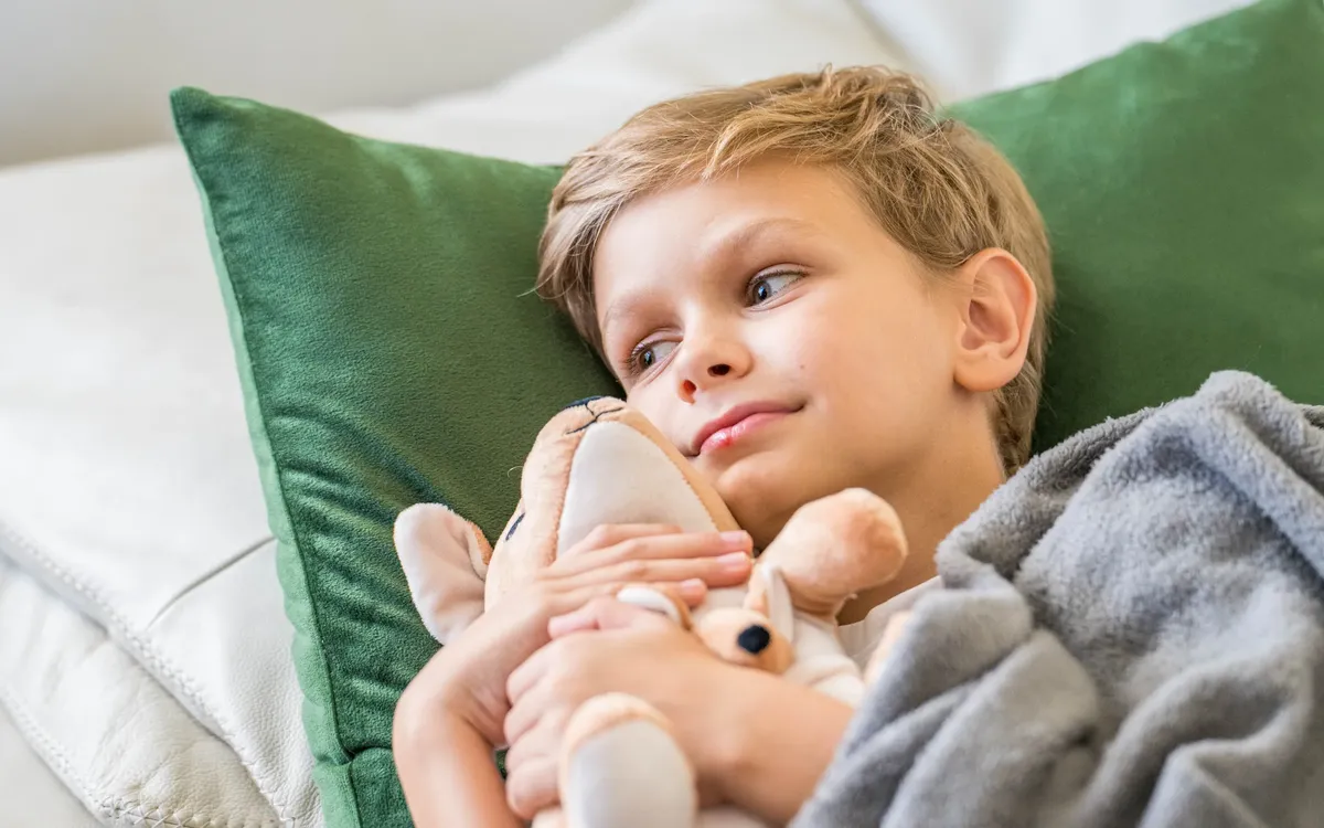 Social Media für APOfit: Ein Bub liegt mit seinem Warmie-Plüschtier auf einem grünen Kissen, zugedeckt mit einer grauen Decke.