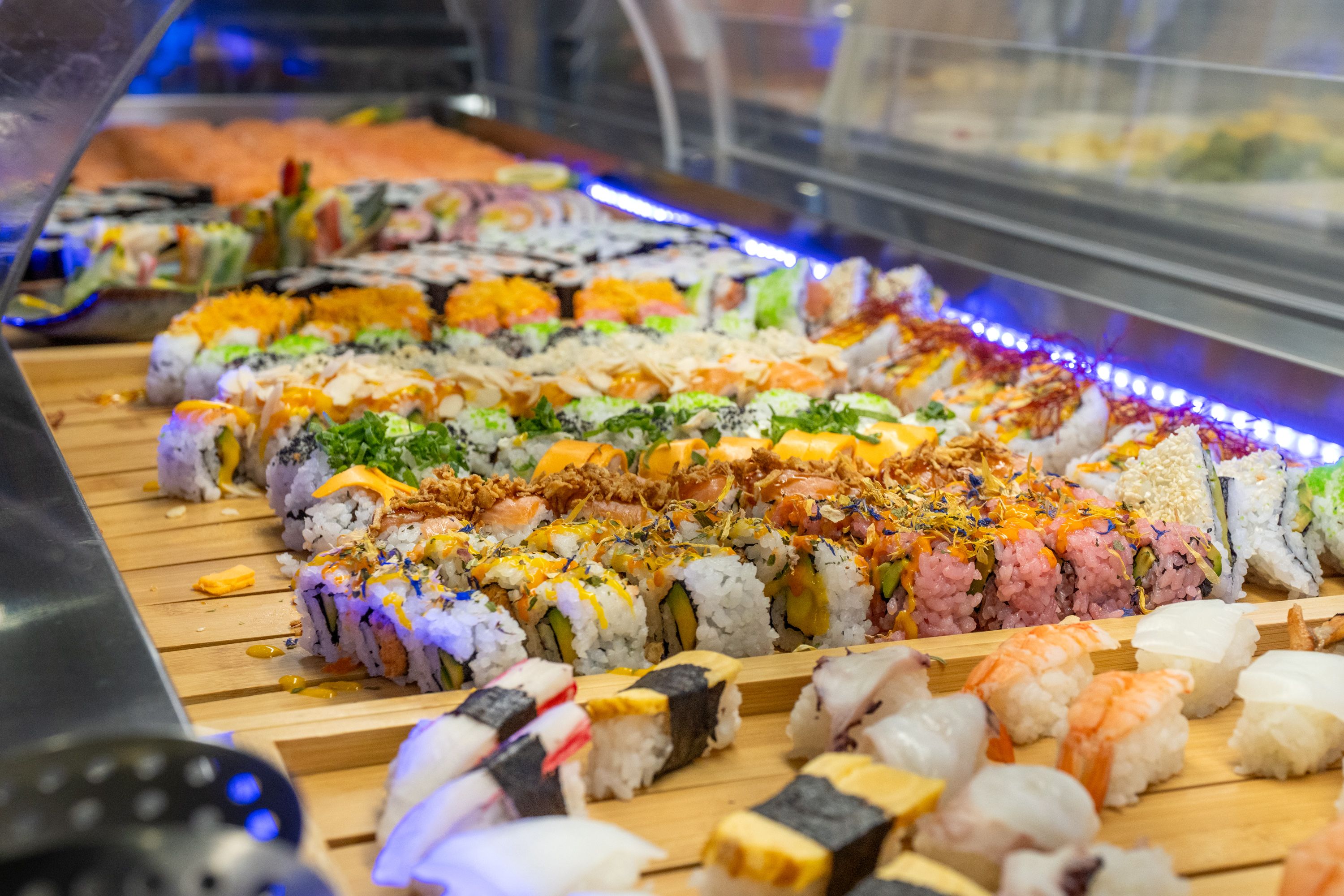 Asia Restaurant YAMI: Eine lange Buffettheke mit Maki-Rollen und Nigiri auf Holzbrettern, darüber ein blaues Lichtband, die Reihe läuft in die Tiefe des Raums.