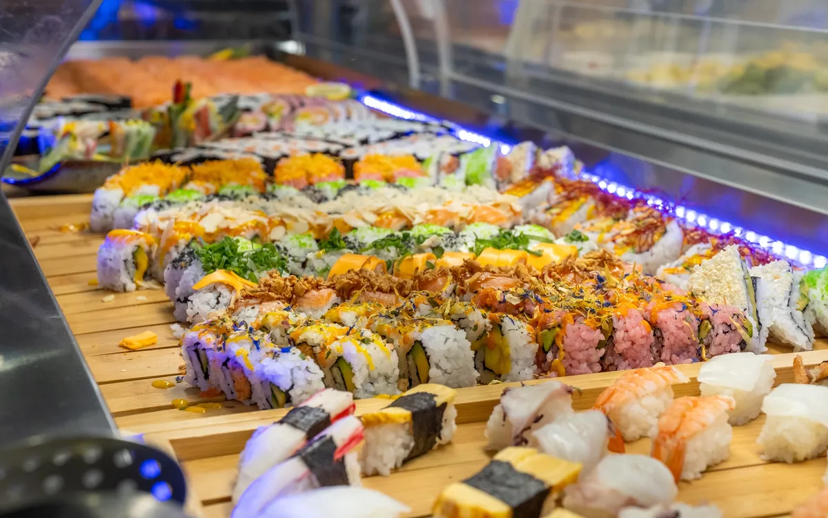 Asia Restaurant YAMI: Eine lange Buffettheke mit Maki-Rollen und Nigiri auf Holzbrettern, darüber ein blaues Lichtband, die Reihe läuft in die Tiefe des Raums.