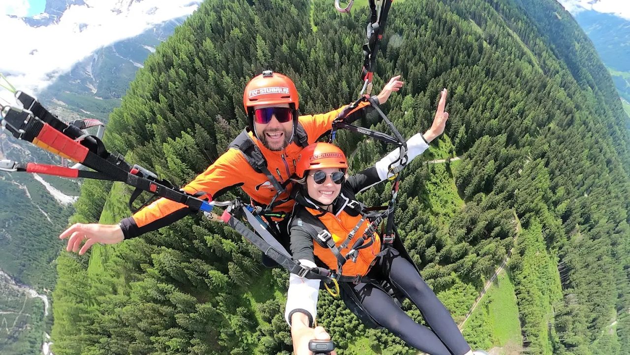 Fly Stubai: Tandemflug im Gleitschirm über bewaldetem Tal, beide Passagiere in orangen Fluganzügen