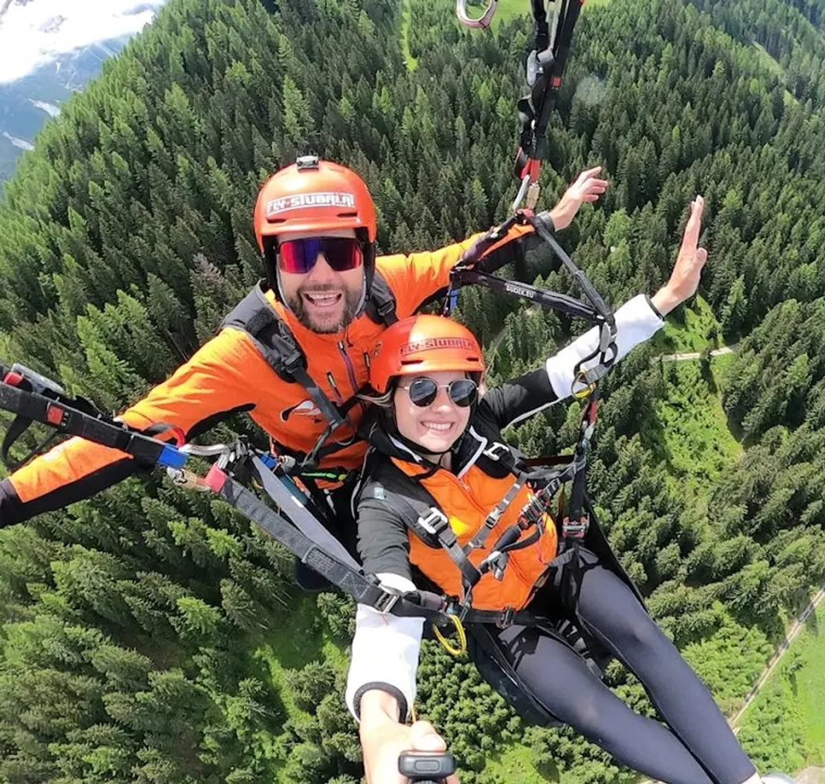 Fly Stubai: Tandemflug im Gleitschirm über bewaldetem Tal, beide Passagiere in orangen Fluganzügen