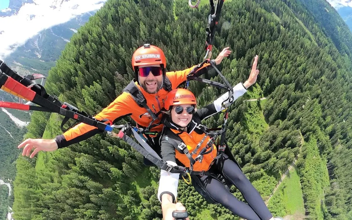 Fly Stubai: Tandemflug im Gleitschirm über bewaldetem Tal, beide Passagiere in orangen Fluganzügen
