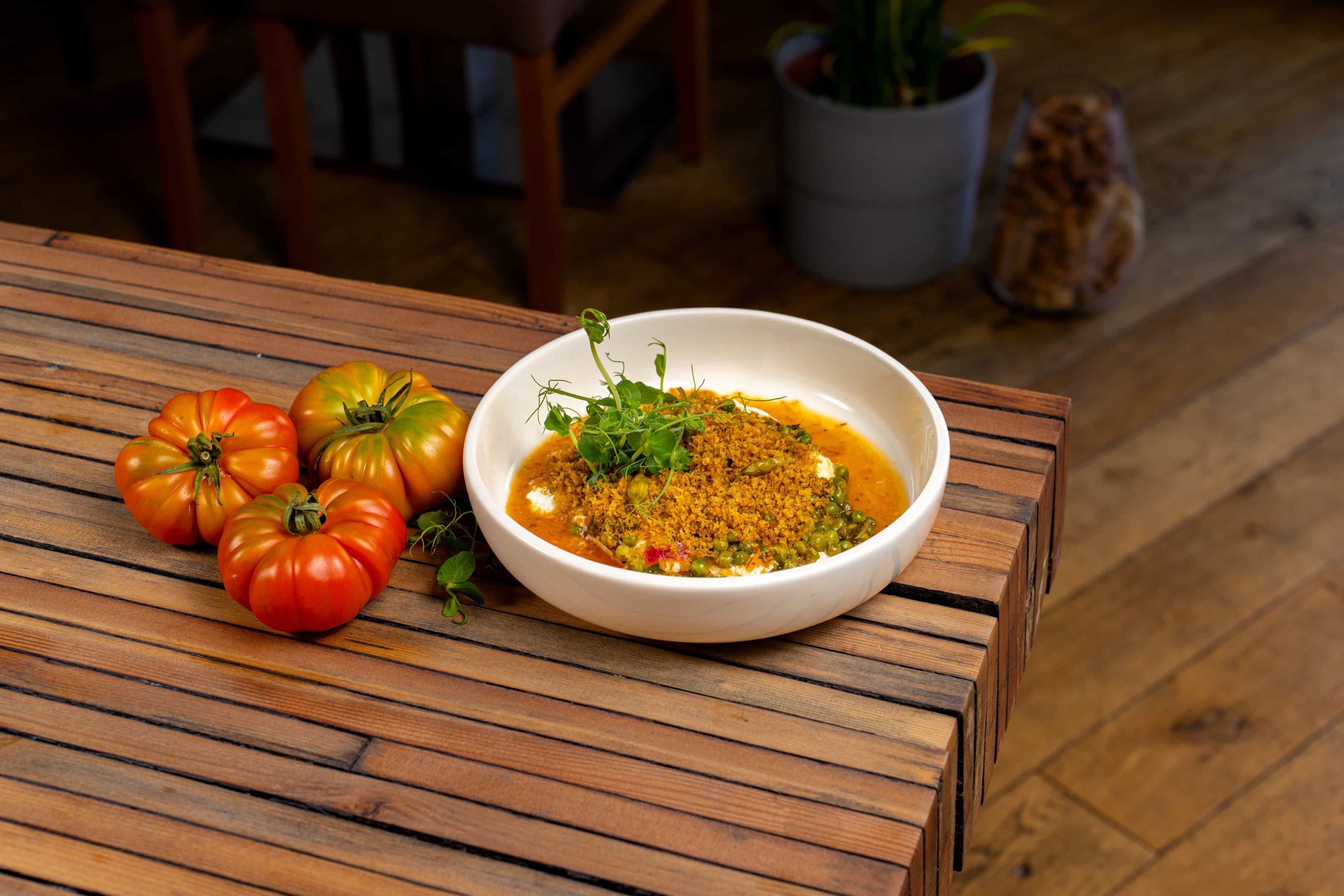 Restaurant BLUM: Eine weisse Schale Curry neben drei Heirloom-Tomaten an der Kante eines Holztischs im Gastraum.