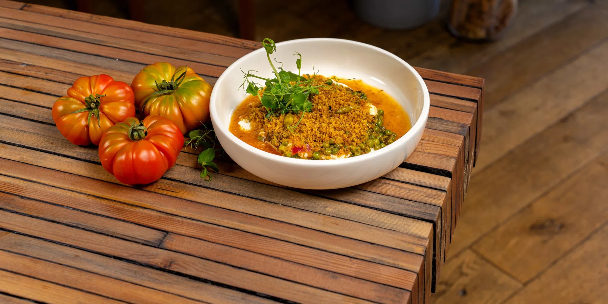 Restaurant BLUM: Eine weisse Schale Curry neben drei Heirloom-Tomaten an der Kante eines Holztischs im Gastraum.