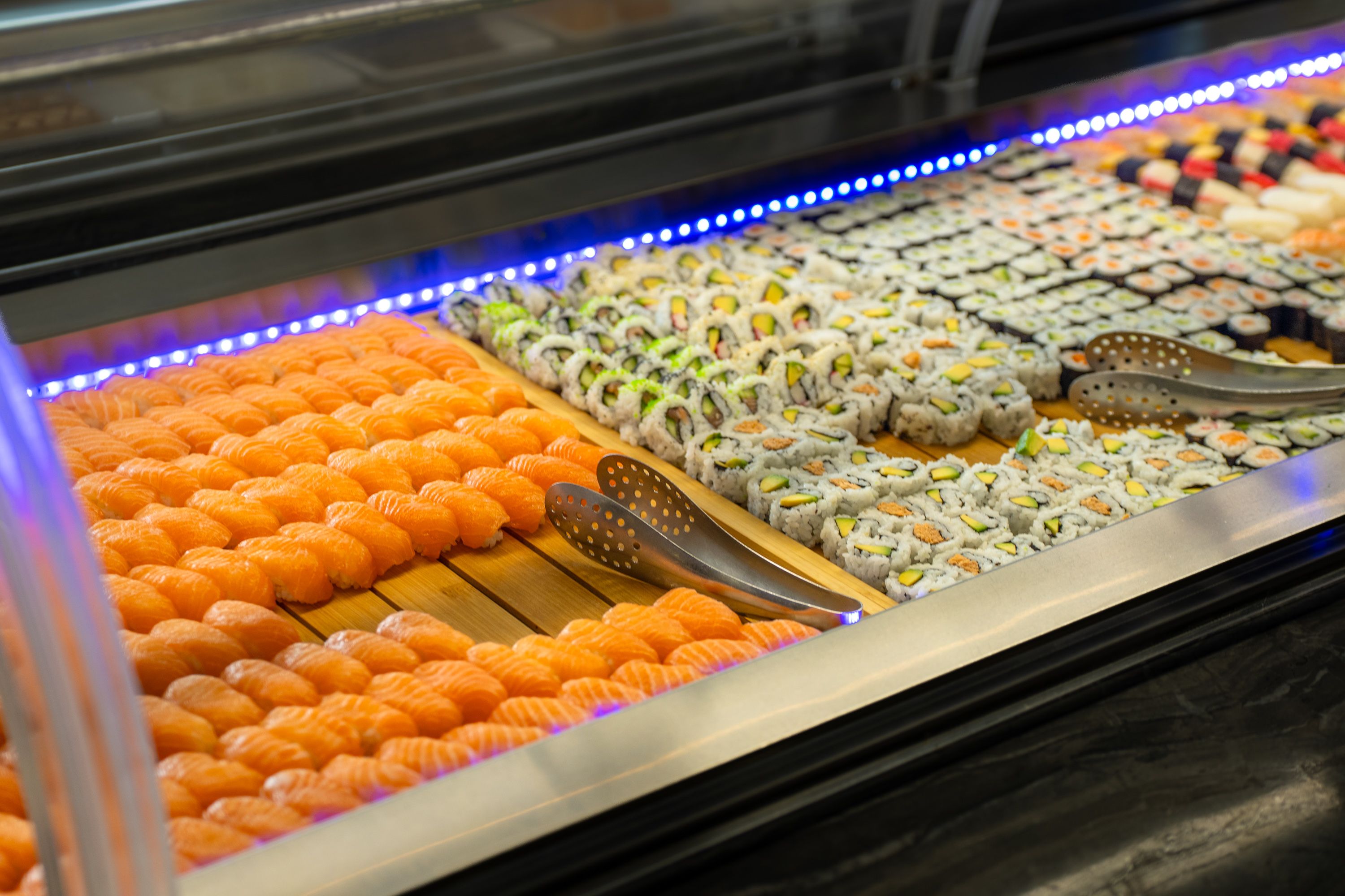 Asia Restaurant YAMI: Reihen Lachs-Nigiri und Maki-Rollen in der Buffetvitrine, darüber das blaue Lichtband der Sushitheke.