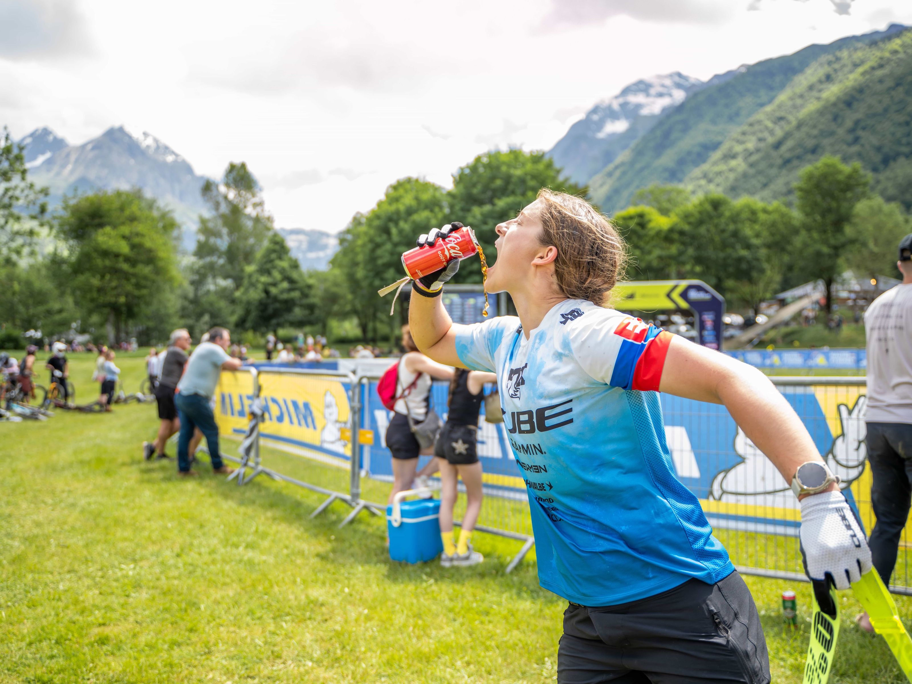 CUBE Actionteam: Mountainbikerin im CUBE-Trikot trinkt im Rennen aus der Flasche, dahinter das Fahrerlager