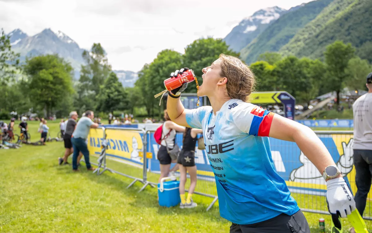 CUBE Actionteam: Mountainbikerin im CUBE-Trikot trinkt im Rennen aus der Flasche, dahinter das Fahrerlager