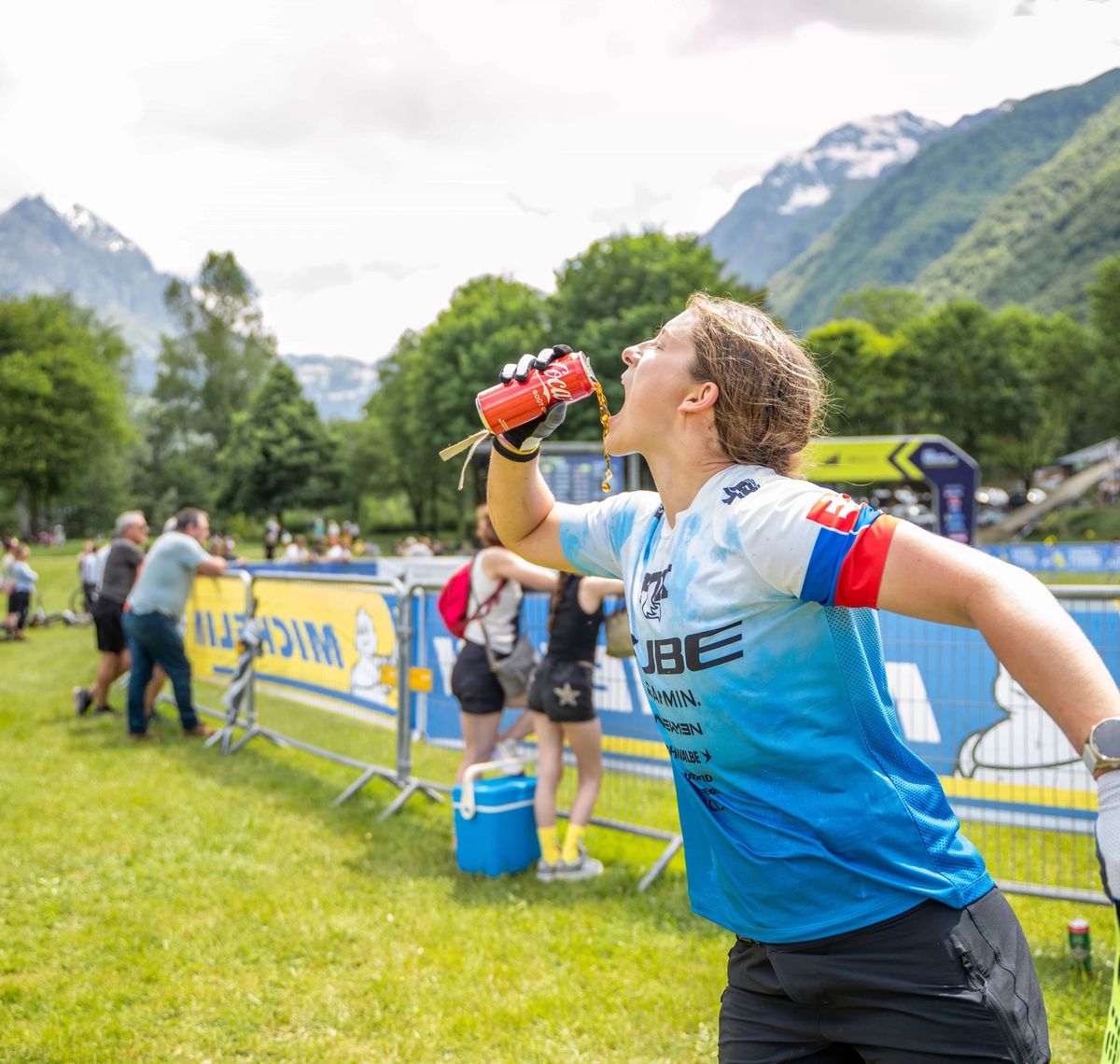 CUBE Actionteam: Mountainbikerin im CUBE-Trikot trinkt im Rennen aus der Flasche, dahinter das Fahrerlager