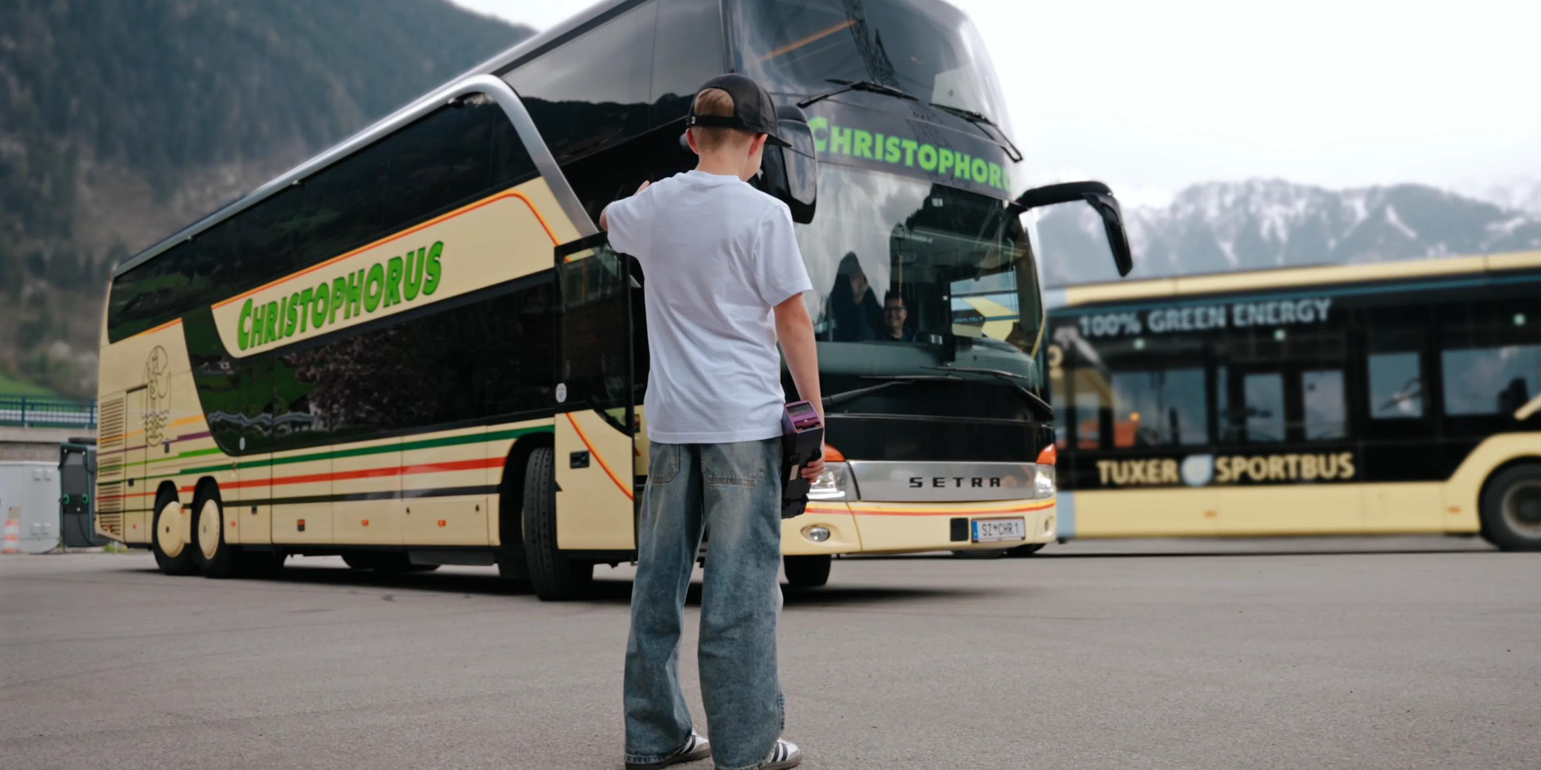 Christophorus Reisen: Ein Junge mit Basecap steht auf einem Parkplatz vor einem Reisebus mit dem Schriftzug Christophorus, dahinter ein zweiter Bus und die bewaldeten Hänge des Zillertals.