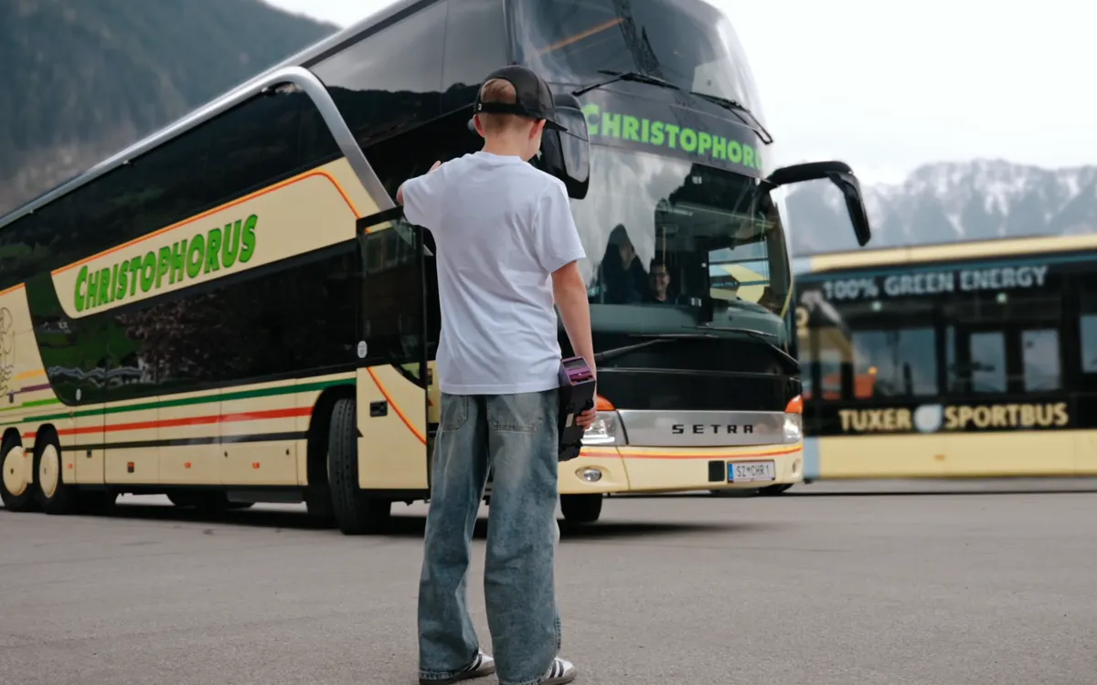 Christophorus Reisen: Ein Junge mit Basecap steht auf einem Parkplatz vor einem Reisebus mit dem Schriftzug Christophorus, dahinter ein zweiter Bus und die bewaldeten Hänge des Zillertals.