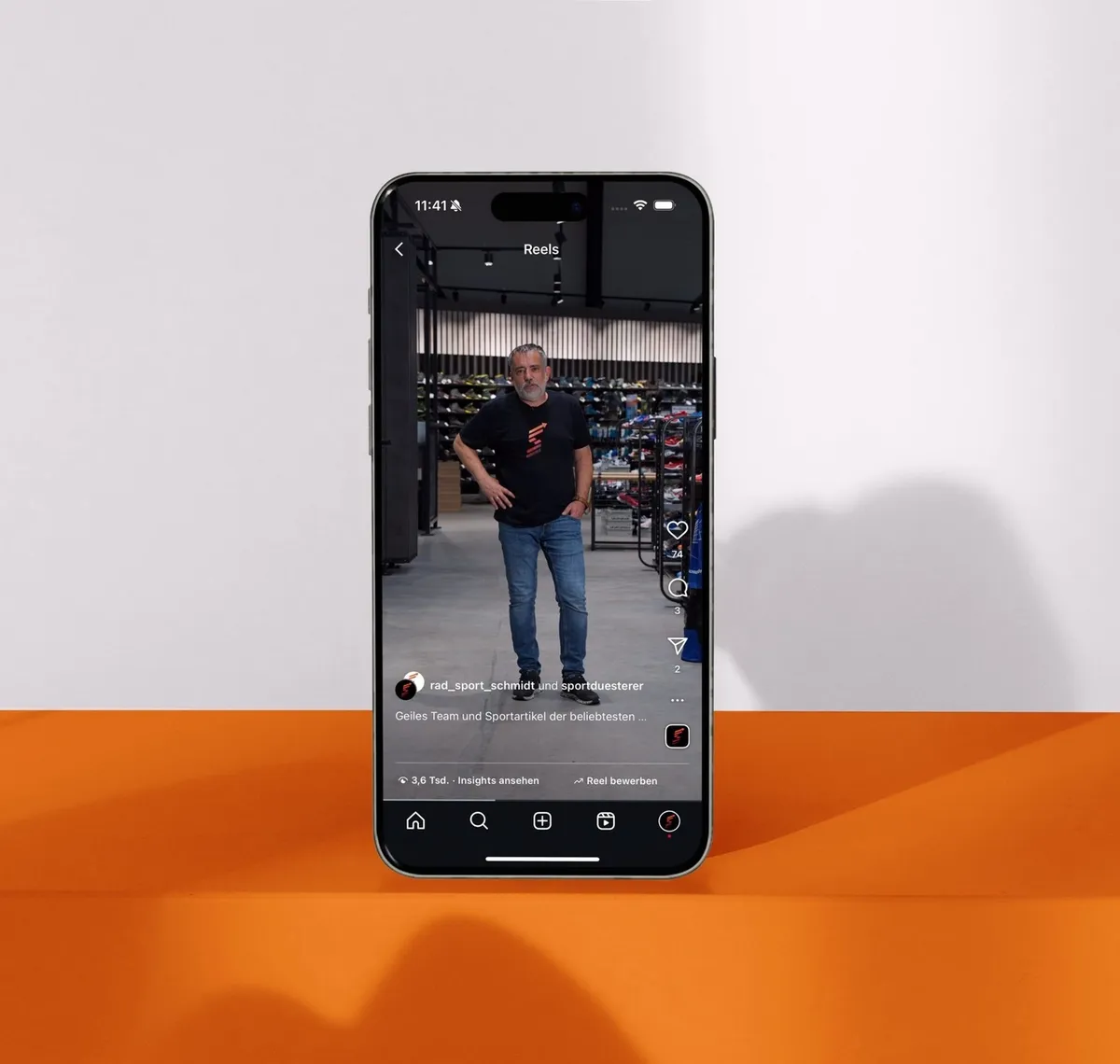 Rad Sport Schmidt: Smartphone über orangem Sockel zeigt ein Recruiting-Reel mit einem Mitarbeiter im Verkaufsraum