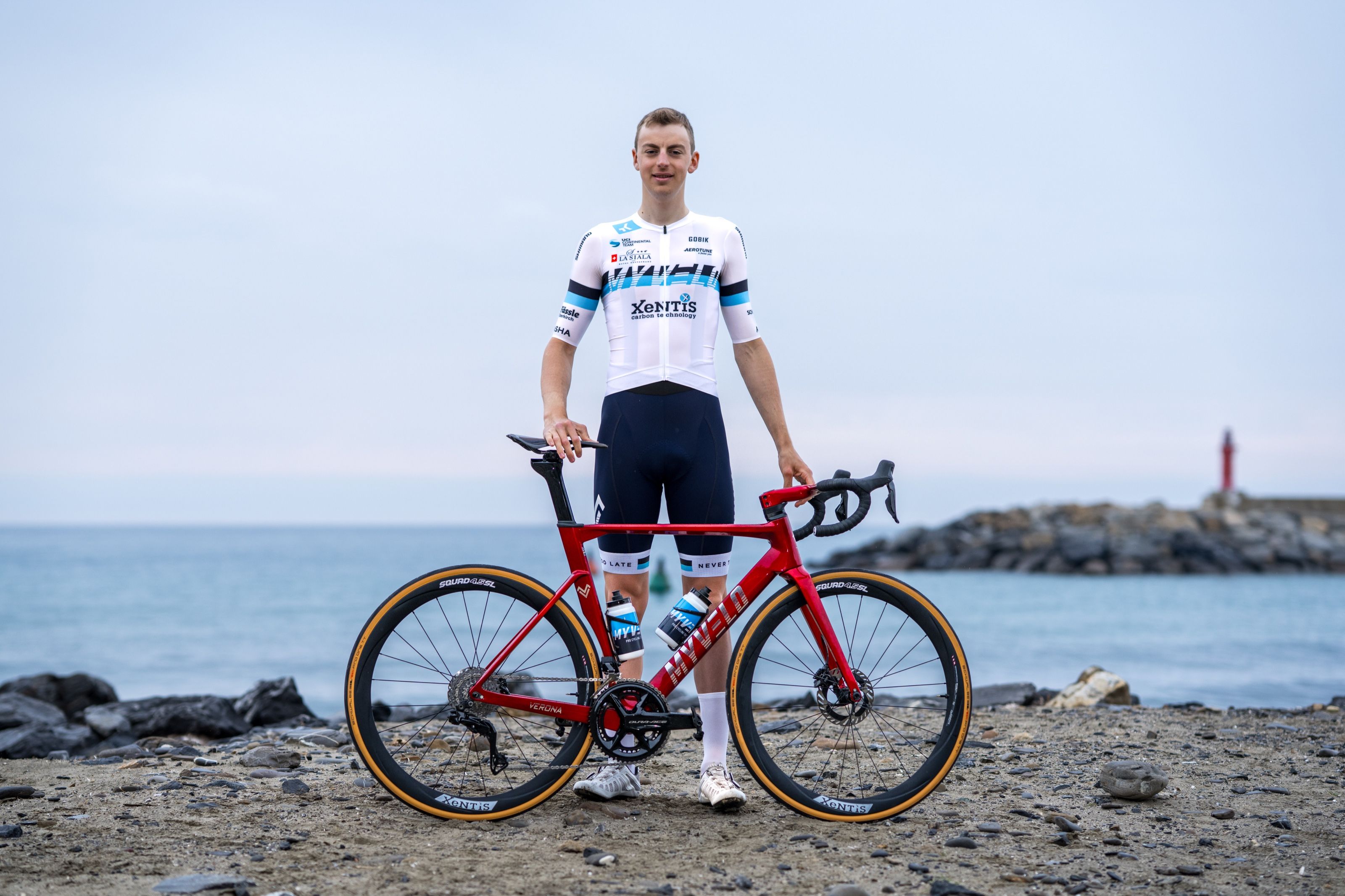 Andrin Züger: Rennradfahrer im weiß-blauen Trikot steht mit seinem roten Rennrad an einem Kiesstrand, dahinter das Meer