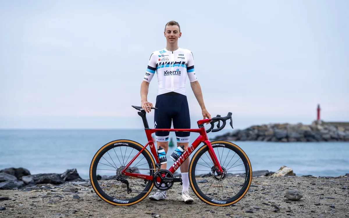 Andrin Züger: Rennradfahrer im weiß-blauen Trikot steht mit seinem roten Rennrad an einem Kiesstrand, dahinter das Meer