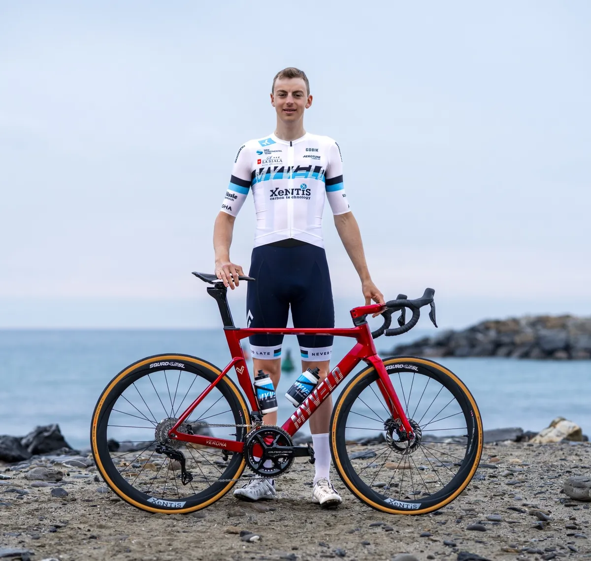 Andrin Züger: Rennradfahrer im weiß-blauen Trikot steht mit seinem roten Rennrad an einem Kiesstrand, dahinter das Meer