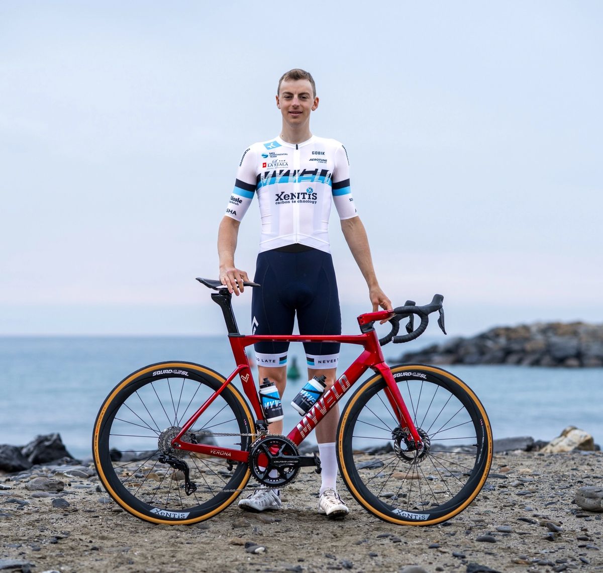 Andrin Züger: Rennradfahrer im weiß-blauen Trikot steht mit seinem roten Rennrad an einem Kiesstrand, dahinter das Meer