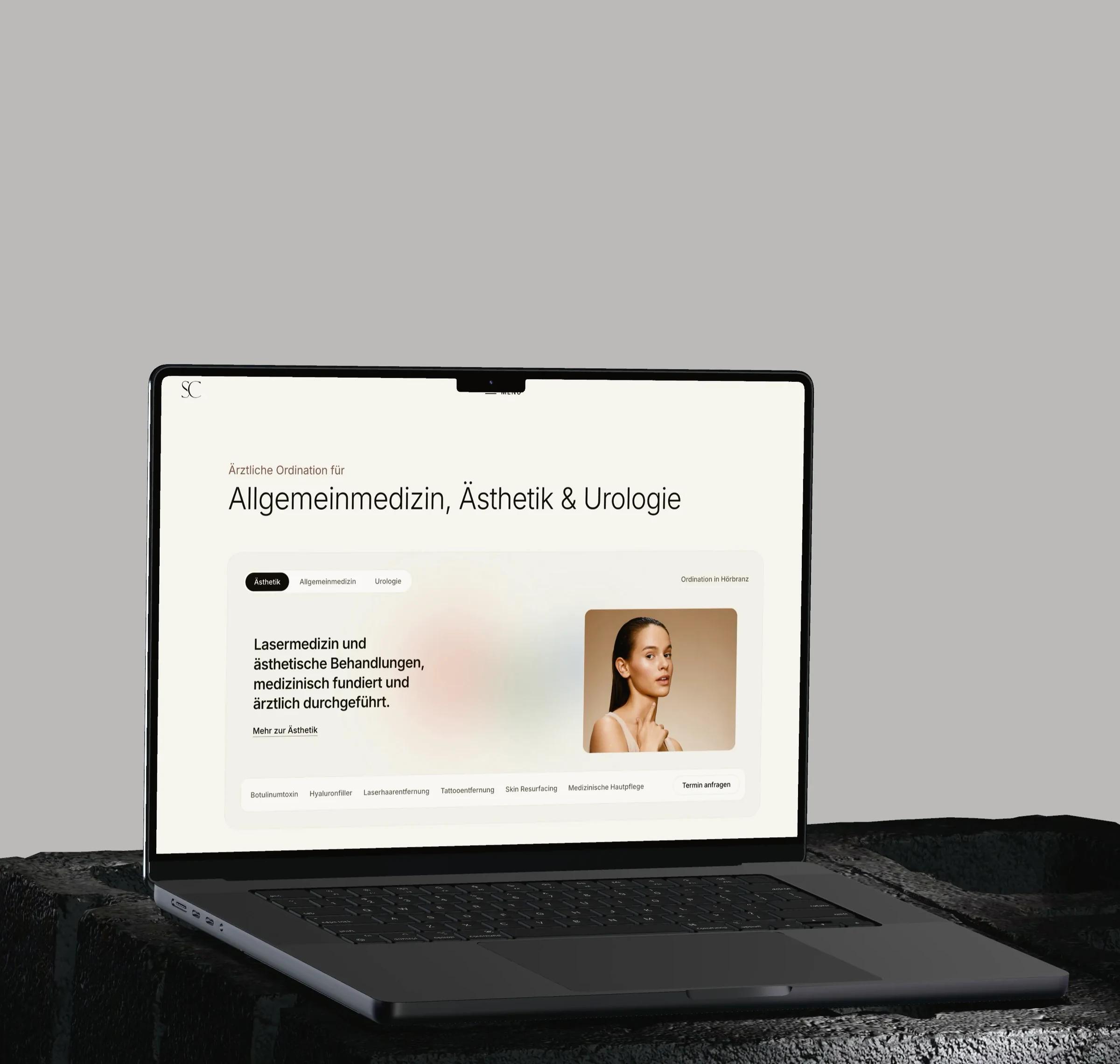 Skinchic.Med: Ein MacBook auf einem dunklen Steinsockel vor hellgrauem Grund zeigt die Startseite von skinchic.at mit den drei Fachbereichen Ästhetik, Allgemeinmedizin und Urologie.