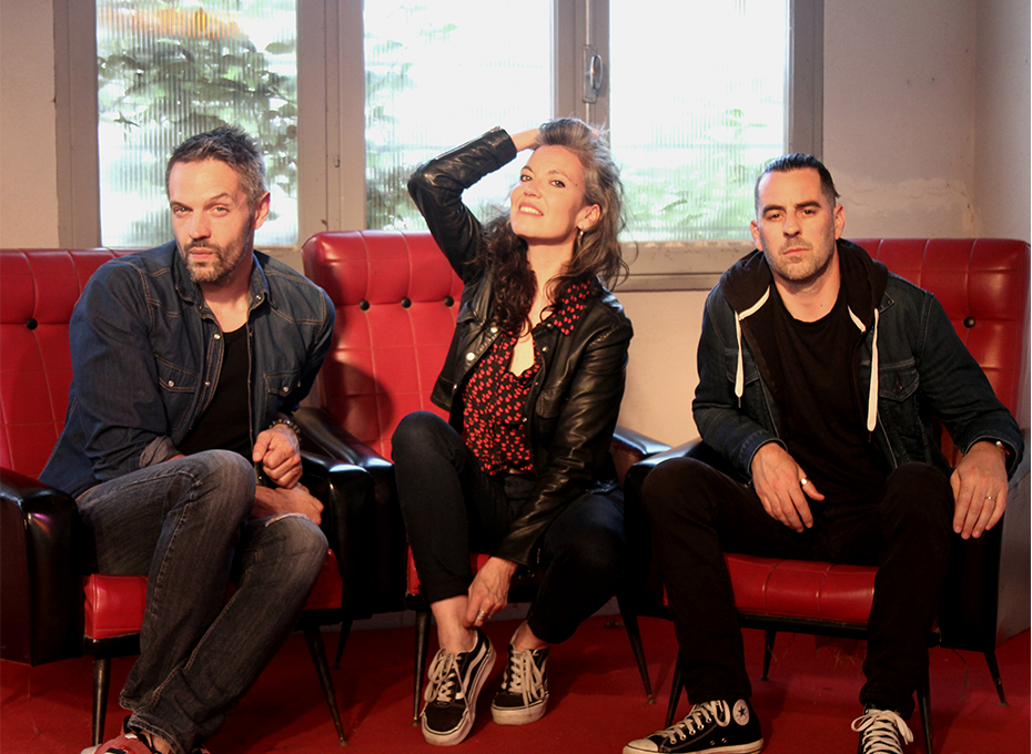 Le groupe Y posant dans un décor aux fauteuils rouges, une chanteuse entre deux musiciens