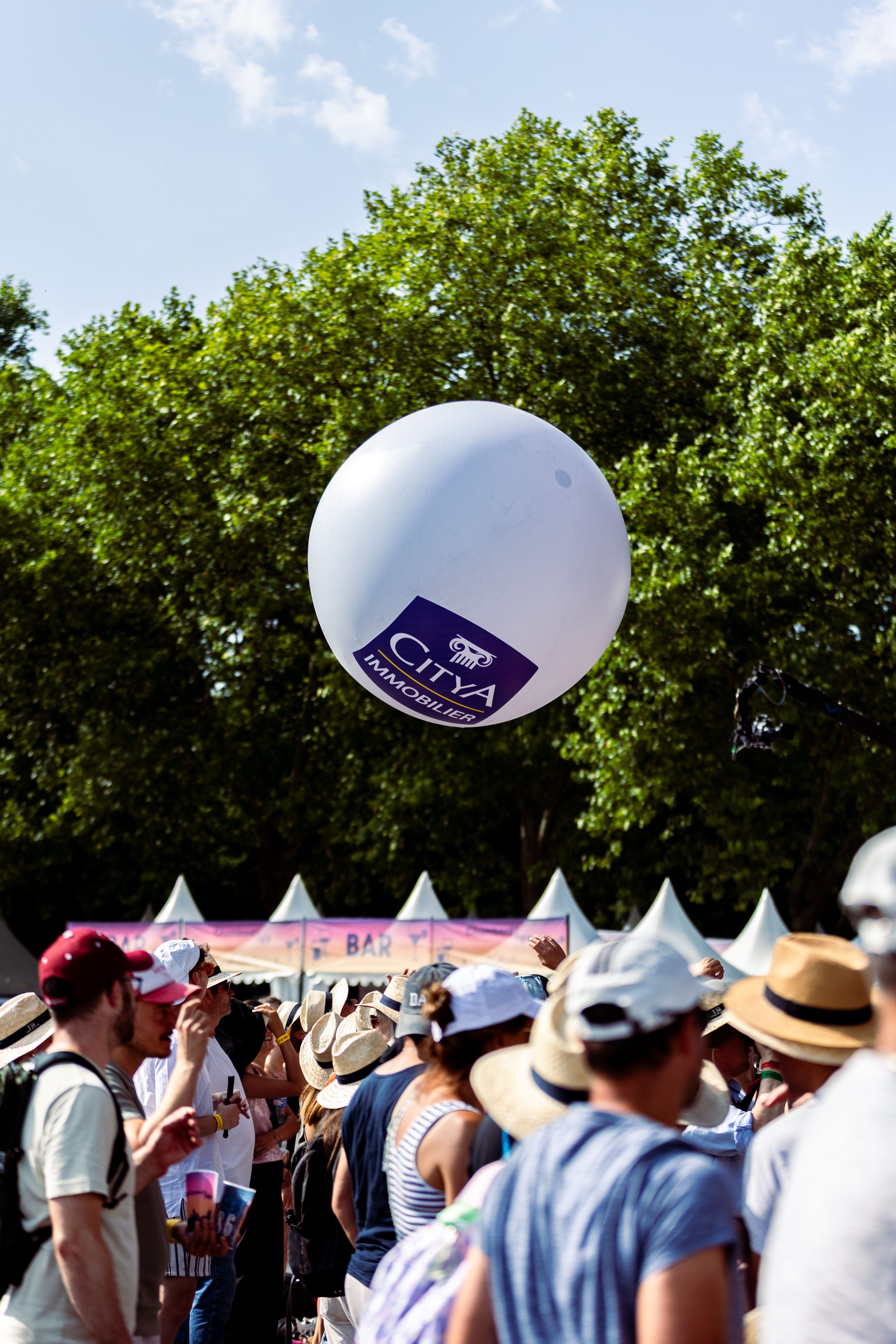 Ballon blanc Citya Immobilier flottant au-dessus du public coiffé de chapeaux de paille, sous les arbres