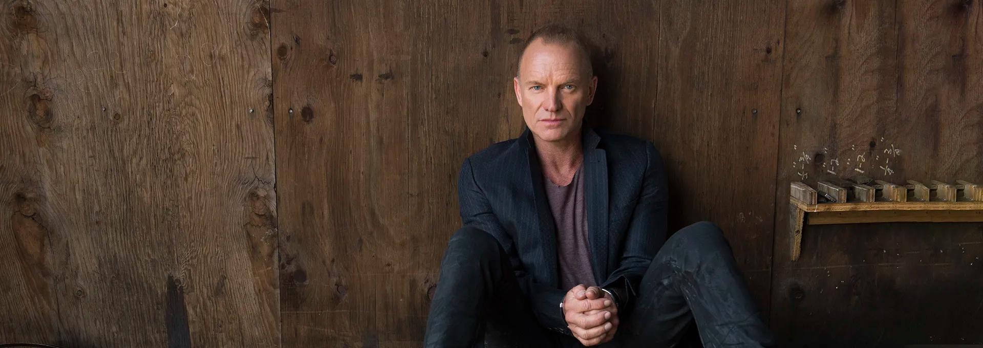 Portrait de Sting assis contre un mur de bois sombre, mains jointes