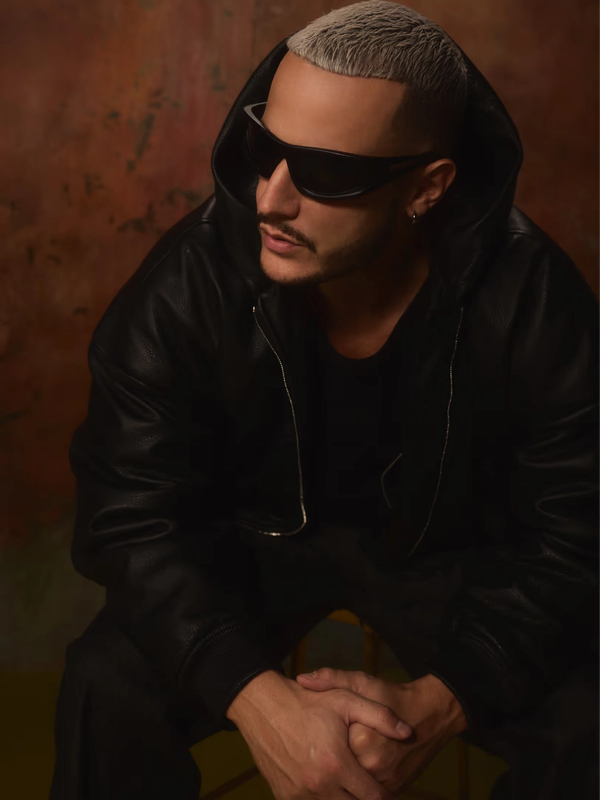DJ Snake en portrait, lunettes noires et veste de cuir à capuche, sur fond cuivré