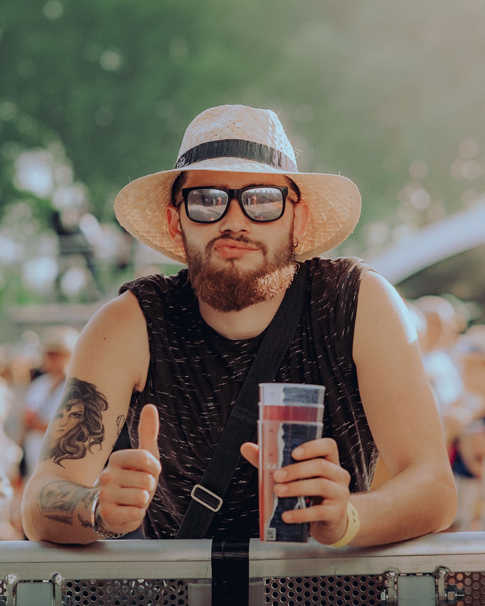 Festivalier en chapeau de paille et lunettes de soleil, pouce levé, un gobelet à la main devant la barrière