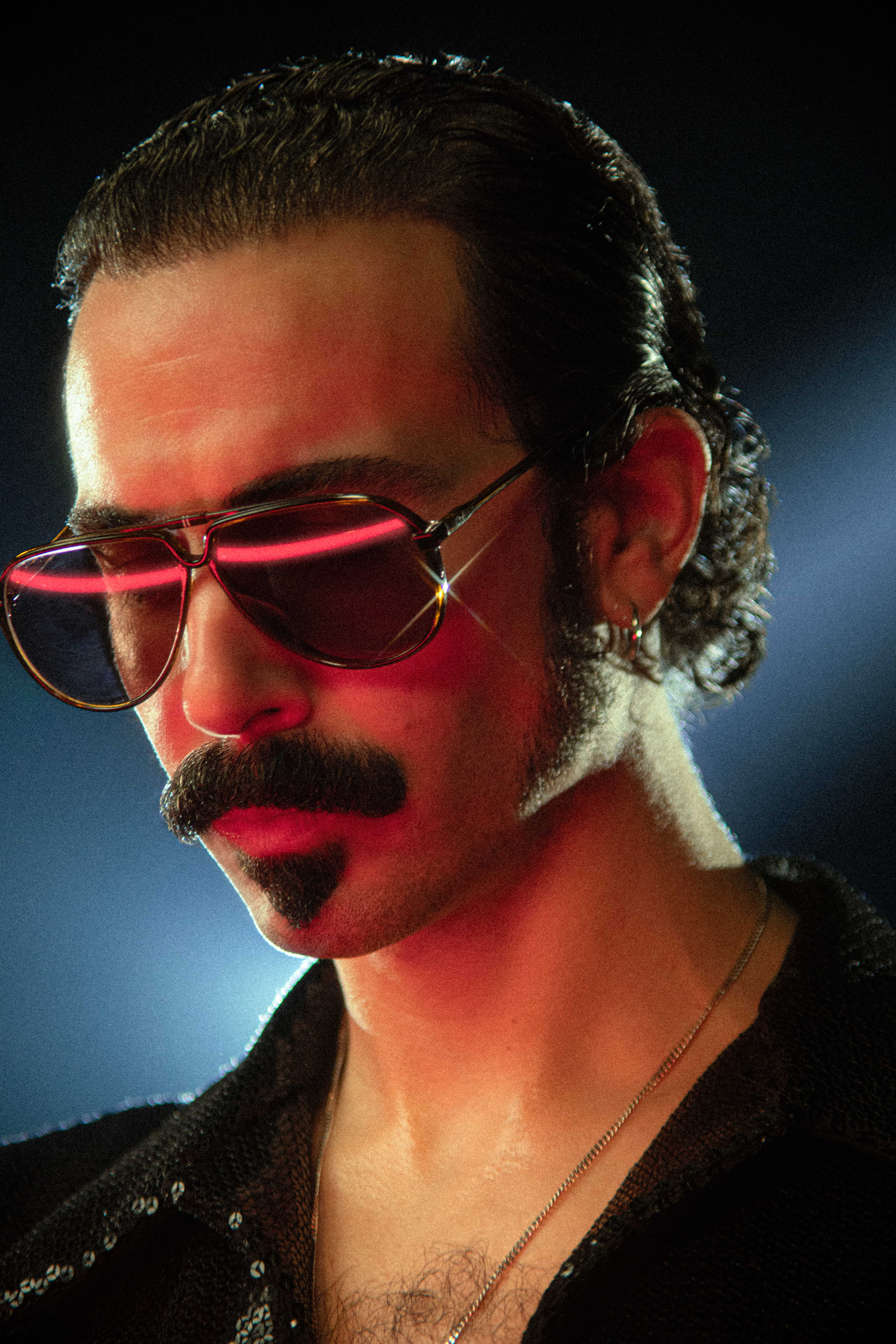 Dabeull en gros plan, lunettes teintées, moustache et lumière rose sur le visage, fond bleu