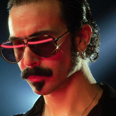 Dabeull en gros plan, lunettes teintées, moustache et lumière rose sur le visage, fond bleu