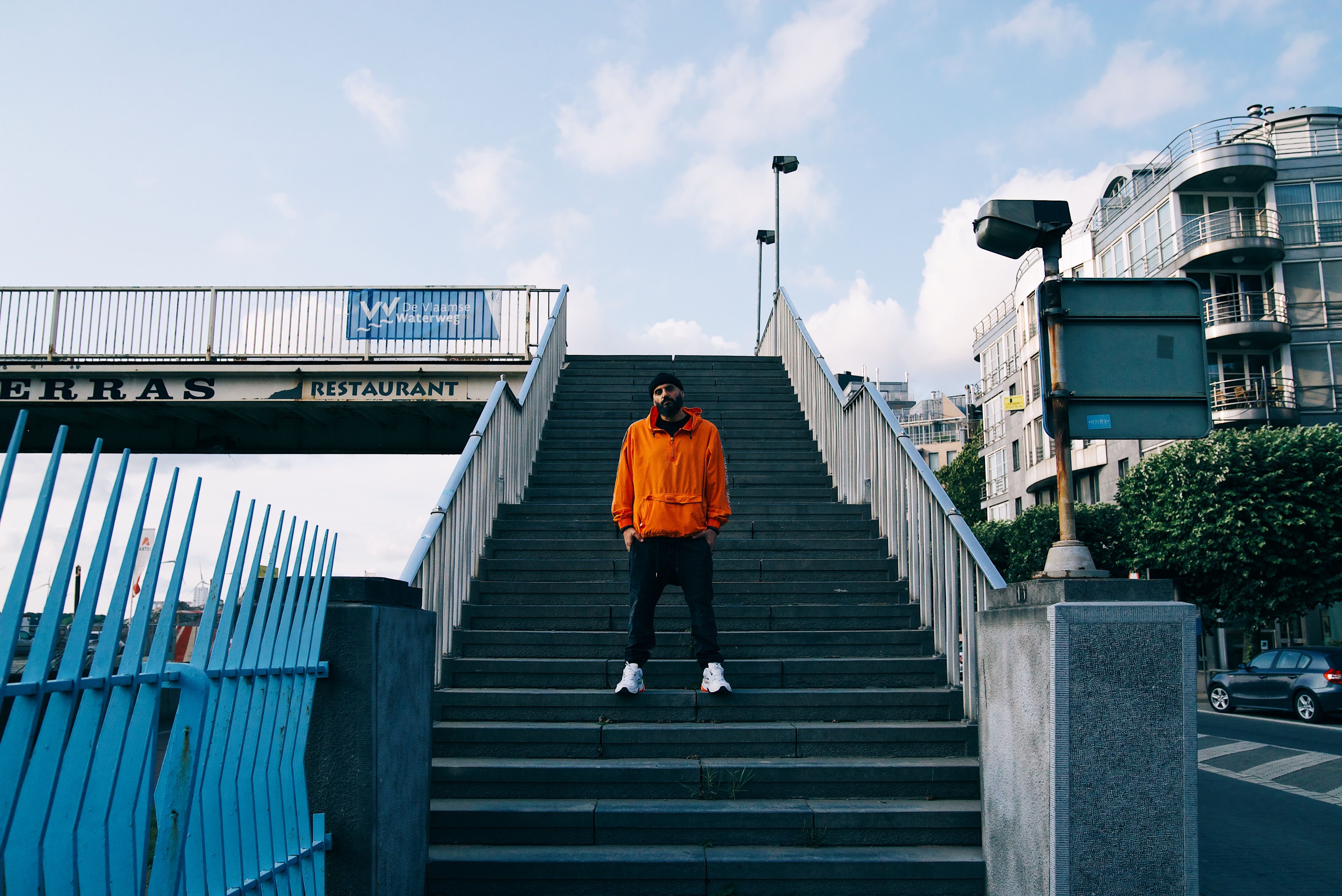 Dany Neville en veste orange en haut d'un escalier, devant une passerelle et des immeubles